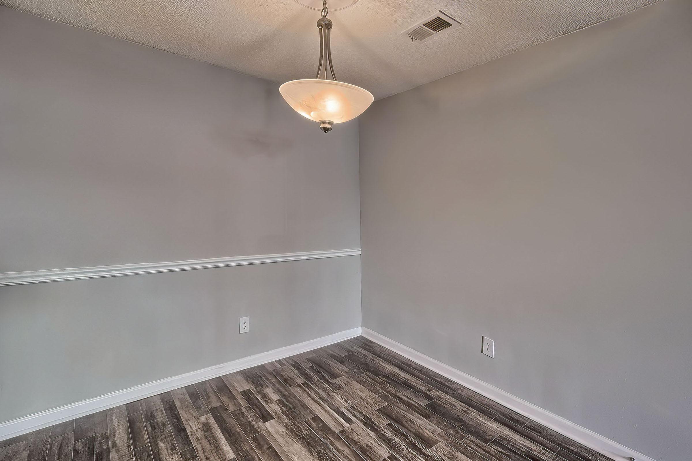 An empty room with gray walls and a pendant light fixture. The floor features wooden planks in a brown hue. There is a white chair rail around the room, and one corner is visible without any furniture or decorations. The overall atmosphere is minimalist and spacious.