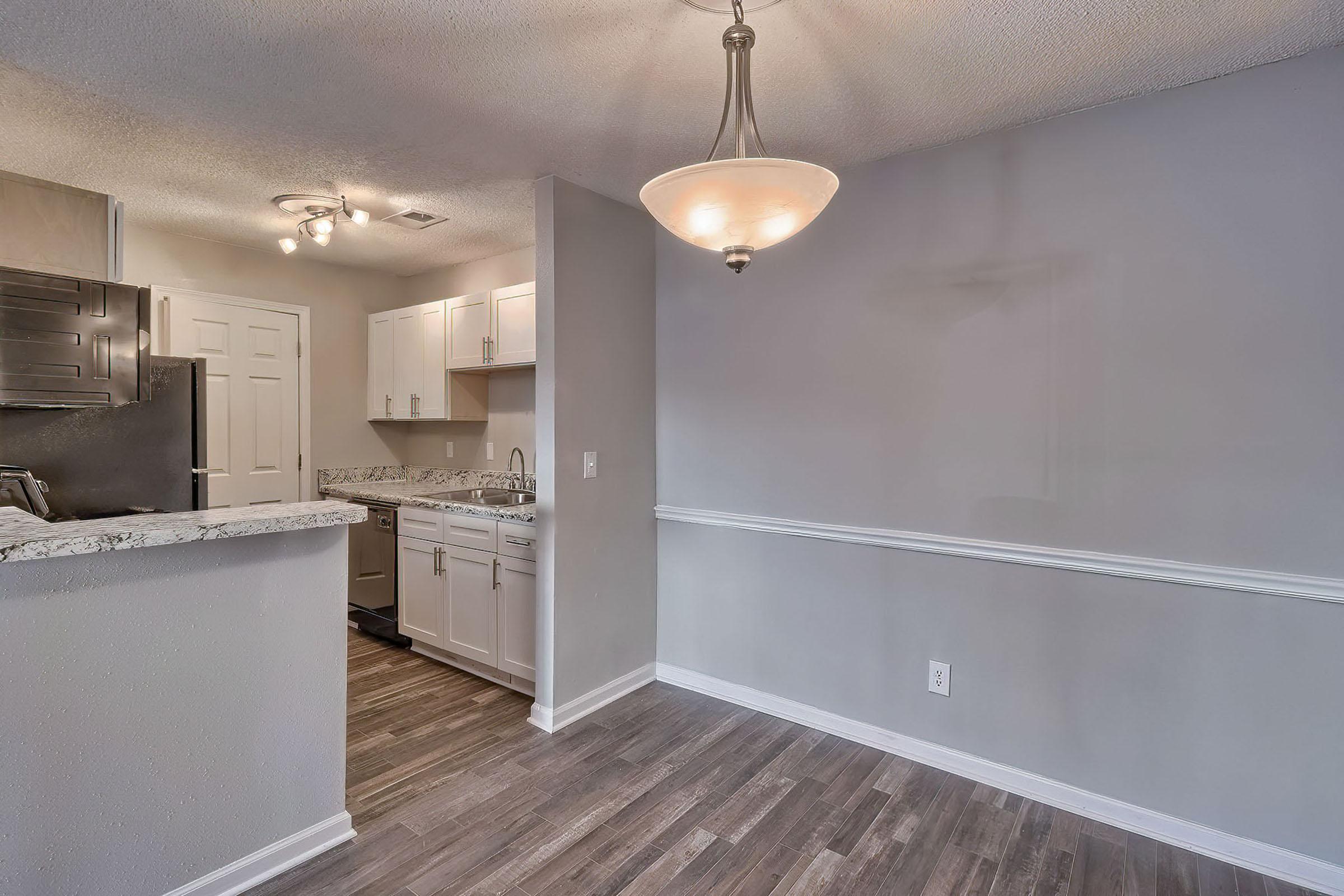 Modern kitchen in an apartment with granite countertops and stainless steel appliances. A dining area is adjacent, featuring a light fixture and neutral-colored walls. The flooring is dark wood, creating a warm atmosphere.