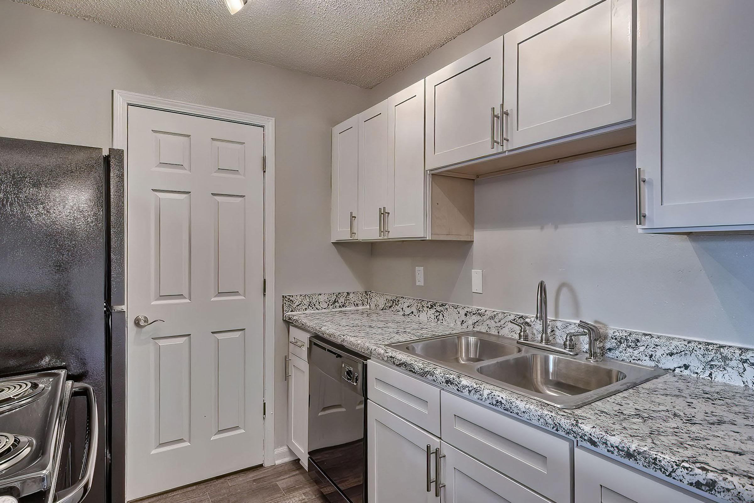 A modern kitchen featuring white cabinets, a granite countertop, a stainless-steel sink, and a dishwasher. An electric stovetop and a black refrigerator are visible. A closed door leads to another area, and the walls are painted a light color, enhancing the bright and clean ambiance.