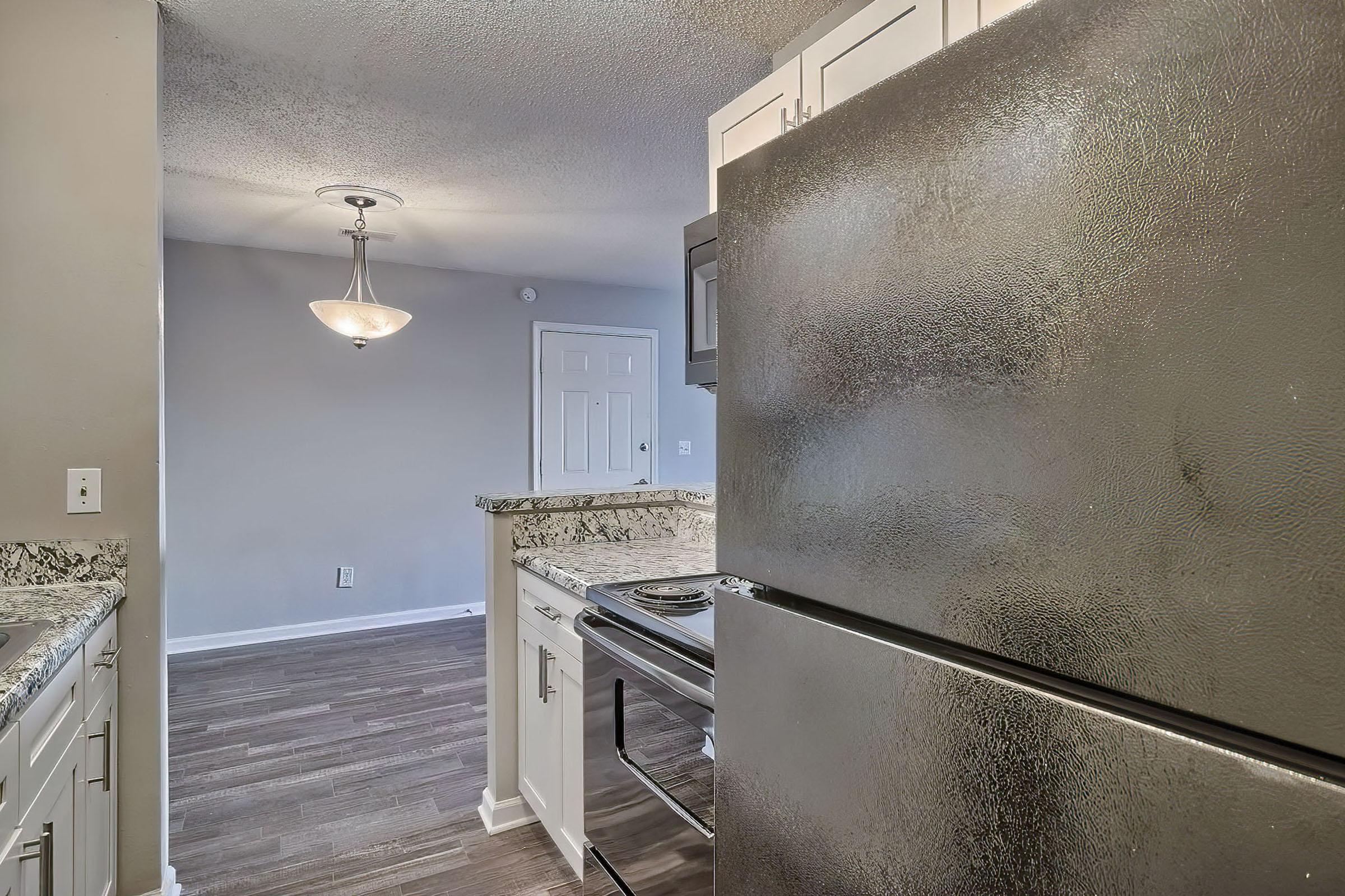 A modern kitchen interior featuring a black refrigerator, white cabinets, and granite countertops. In the background, there's a doorway leading to another room, illuminated by a pendant light hanging from the ceiling. The floor is finished with dark wooden planks, creating a sleek and contemporary look.