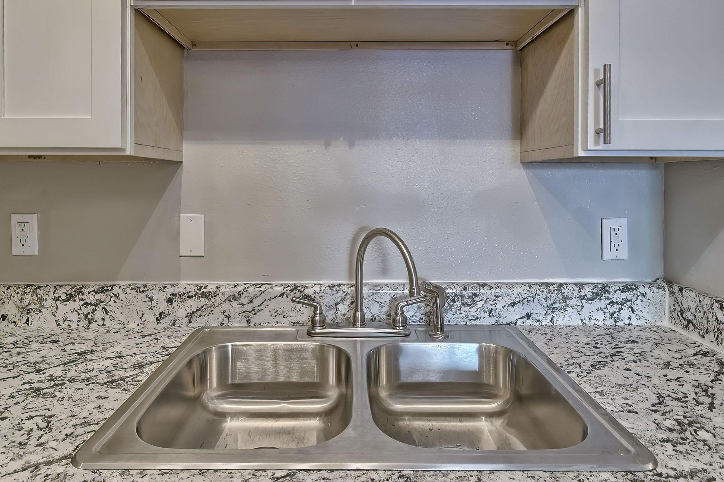 A modern kitchen sink with a double basin, featuring a polished stainless steel finish. The sink is installed on a speckled granite countertop with light and dark gray tones. Above the sink, there are white cabinets and a plain gray wall, emphasizing a clean and contemporary design.