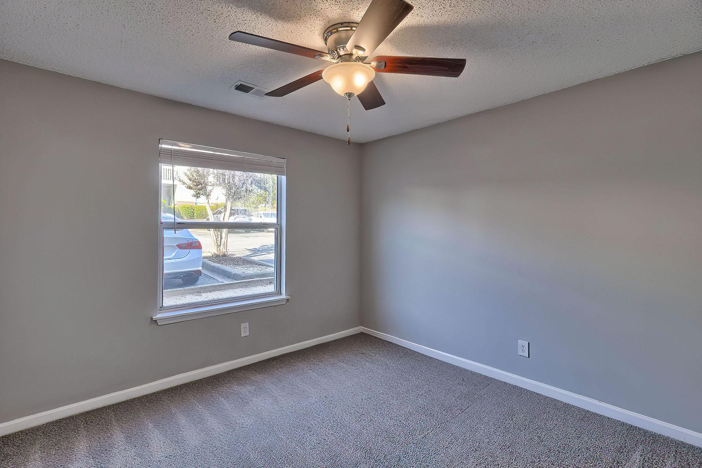 A spacious, empty room featuring light gray walls and a ceiling fan with wooden blades. A window allows natural light to enter, revealing a view of a parked car outside. The floor is covered with soft, beige carpeting. The atmosphere is bright and inviting, perfect for personalization.