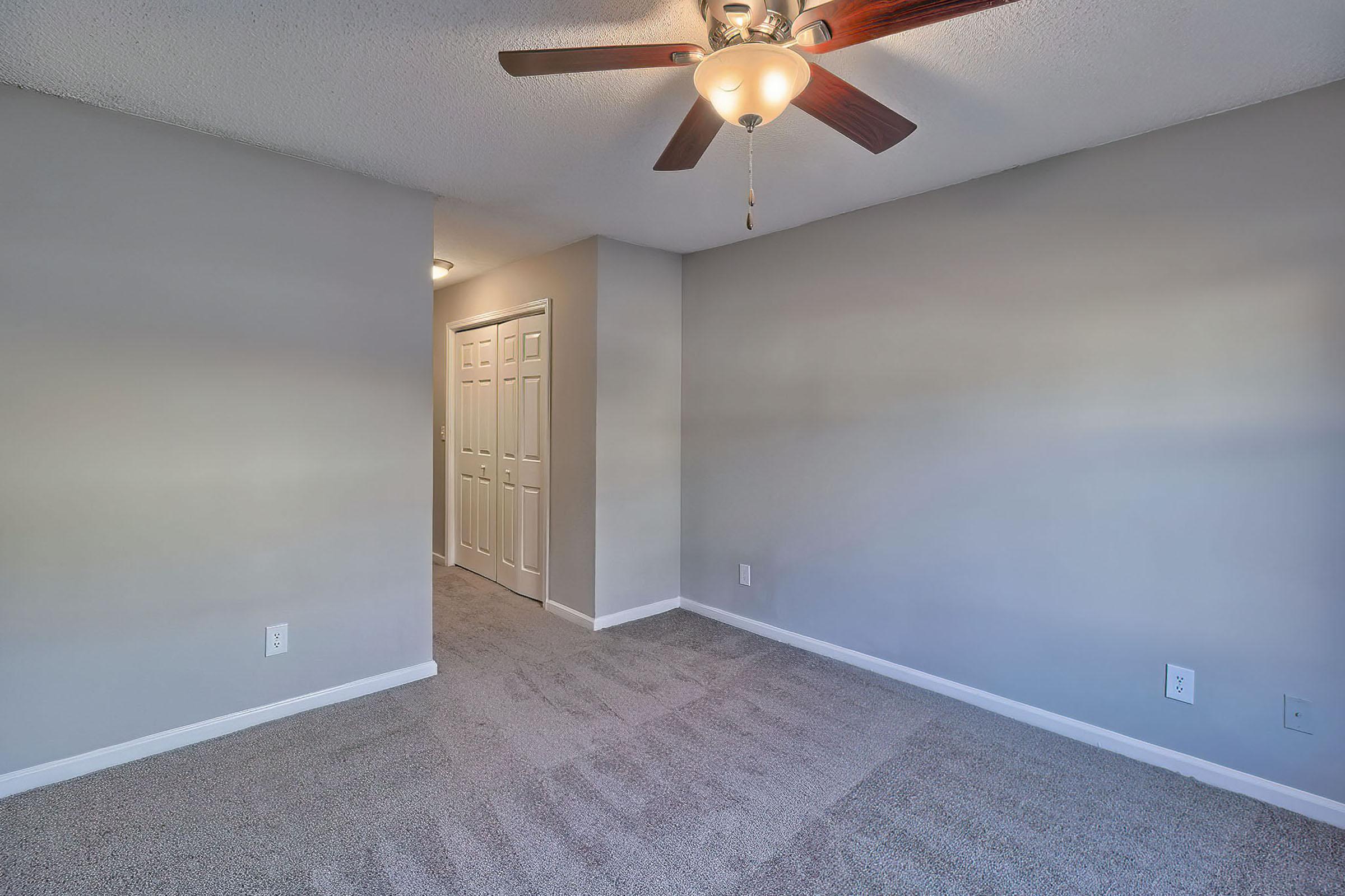 A vacant room with light gray walls and beige carpet. A ceiling fan with wooden blades hangs above. A doorway leads to a hallway with white closet doors. The space is well-lit, creating a welcoming atmosphere, perfect for personalization and decorating.