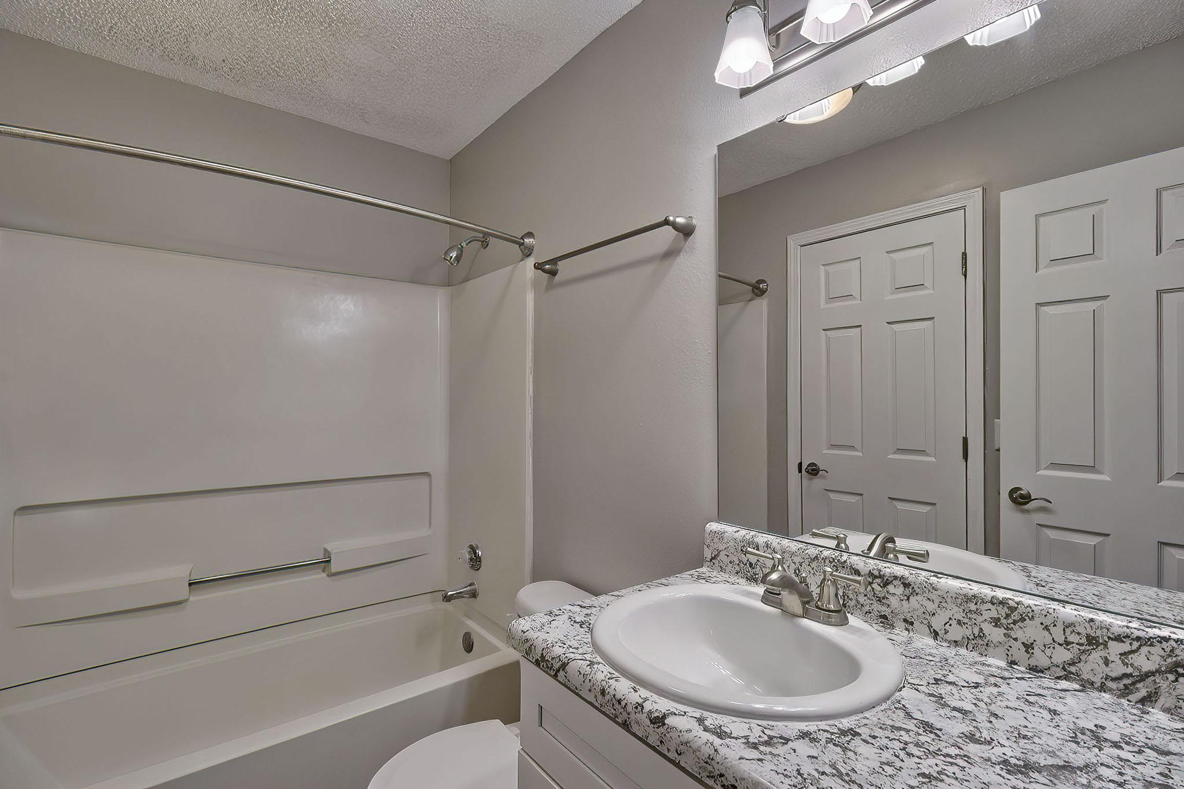 A modern bathroom featuring a white bathtub, a granite countertop with a sink, and a large mirror. The walls are painted in a neutral color, and there is a towel bar mounted on the wall. A door and reflective surfaces create a sense of space and light. The lighting fixtures are above the mirror.