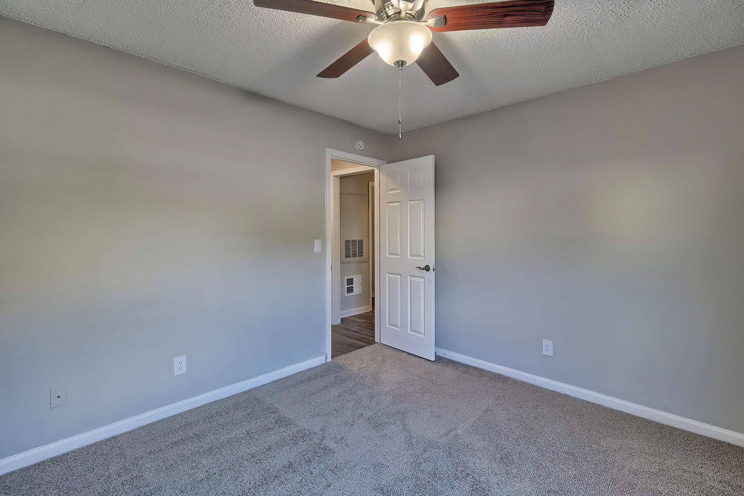 Empty room with gray walls and beige carpet. A ceiling fan with wooden blades is mounted in the center. A door is slightly open, leading to another space. The lighting is bright, creating an inviting atmosphere. No furniture is present in the room.