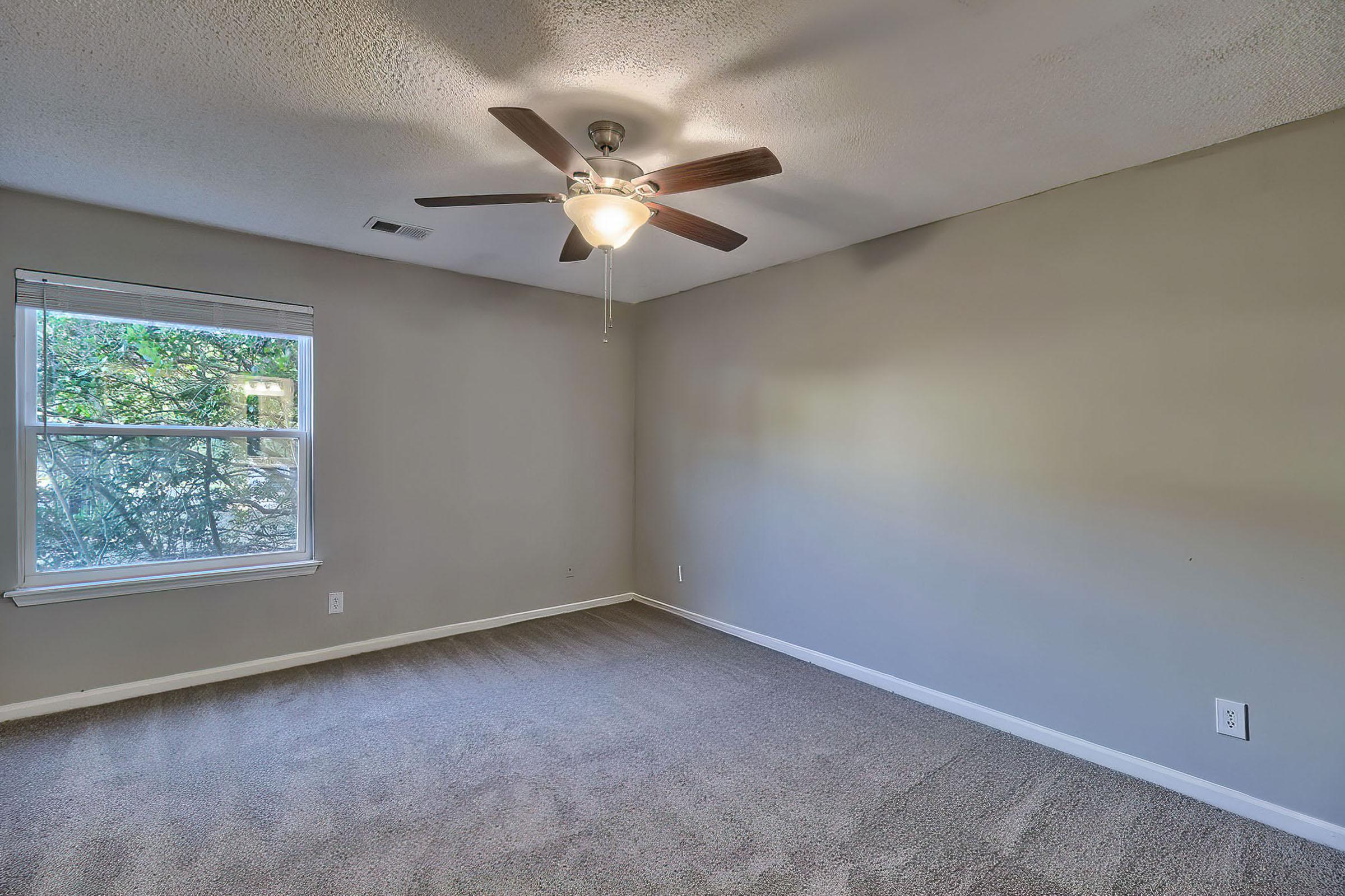 A spacious, empty room with light gray walls and a ceiling fan. The room features a large window providing natural light, with a view of greenery outside. The floor is covered in soft, tan carpet, creating a cozy atmosphere. Ideal for various uses such as a bedroom or office space.