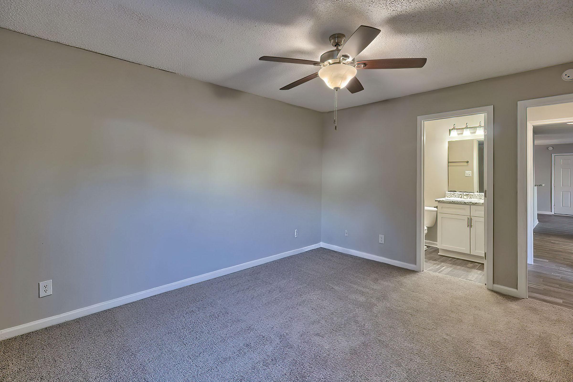 A spacious, well-lit room with a ceiling fan, light gray walls, and beige carpet. A doorway leads to a bathroom with modern fixtures. The room appears fresh and clean, ideal for various uses such as a bedroom or office. Natural light flows in, enhancing the inviting atmosphere.