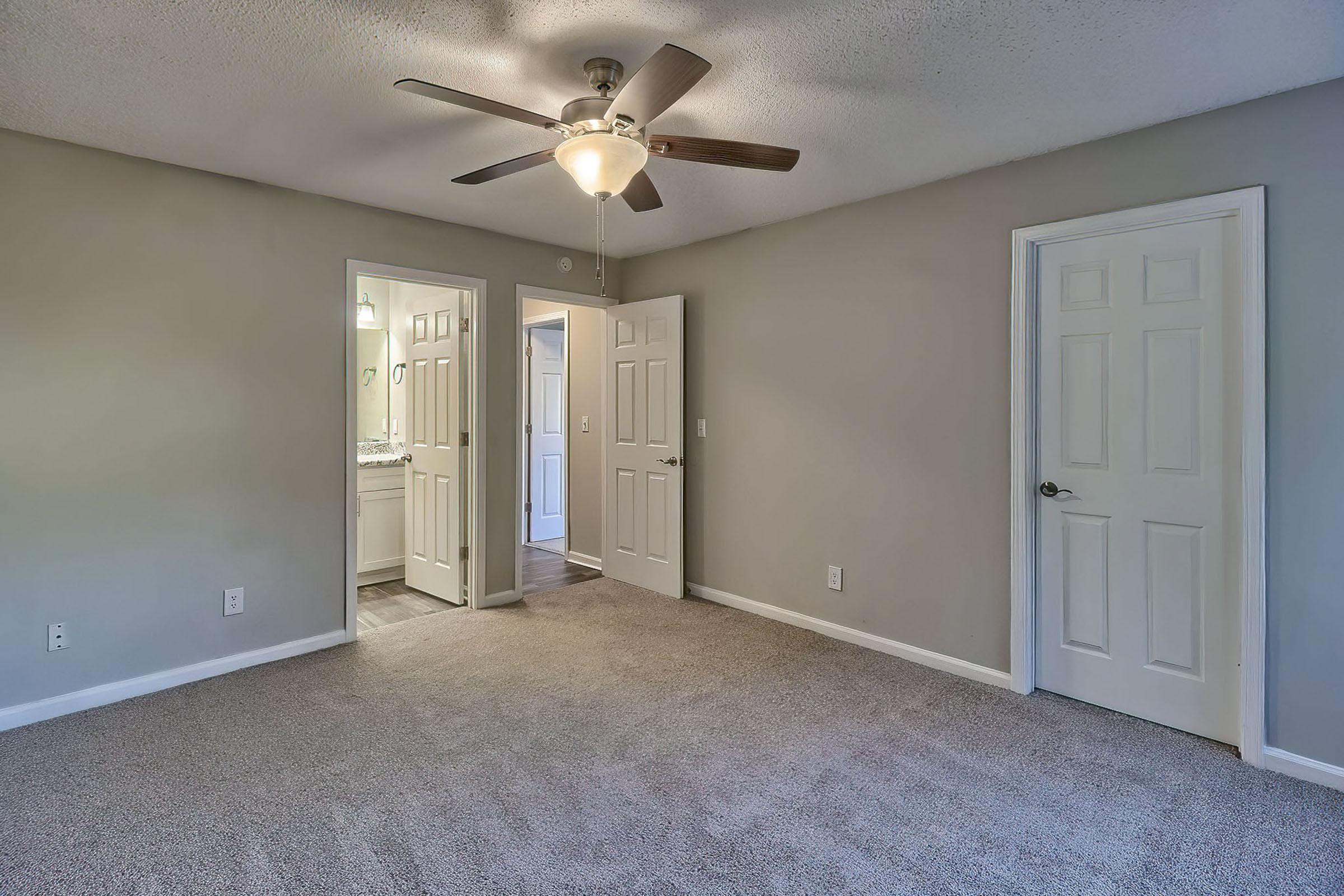 A well-lit room featuring a ceiling fan, beige carpet, and light gray walls. Two closed doors lead to a bathroom and another space. The room is minimalistic with natural light filtering through the doorway, creating a warm and inviting atmosphere.