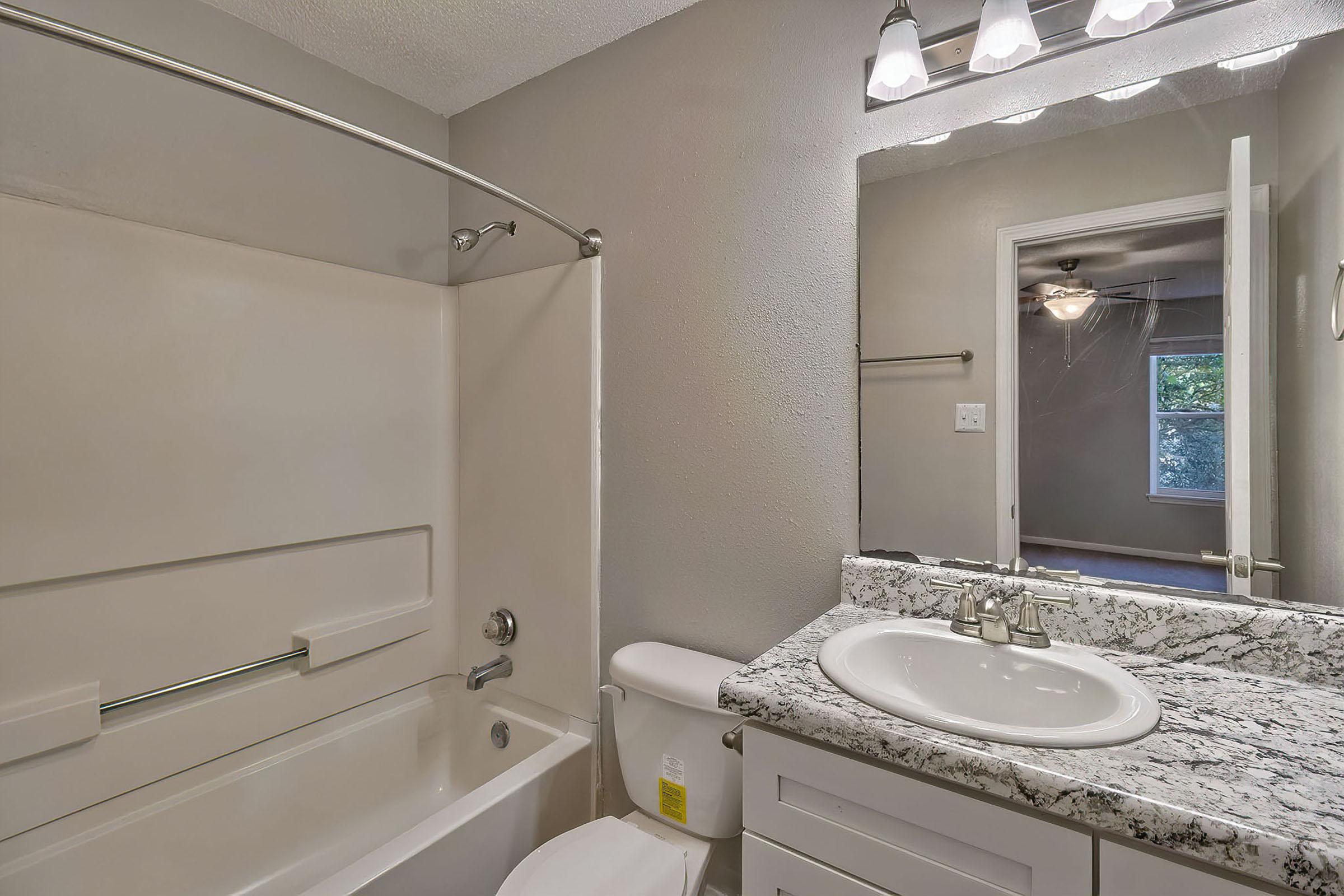 A modern bathroom featuring a combination bathtub and shower with a sliding glass door, vanity with a marble countertop and sink, and a toilet. The walls are painted a light gray, and there is ample natural light coming from an adjacent room with an open door. Ceiling lights provide additional illumination.