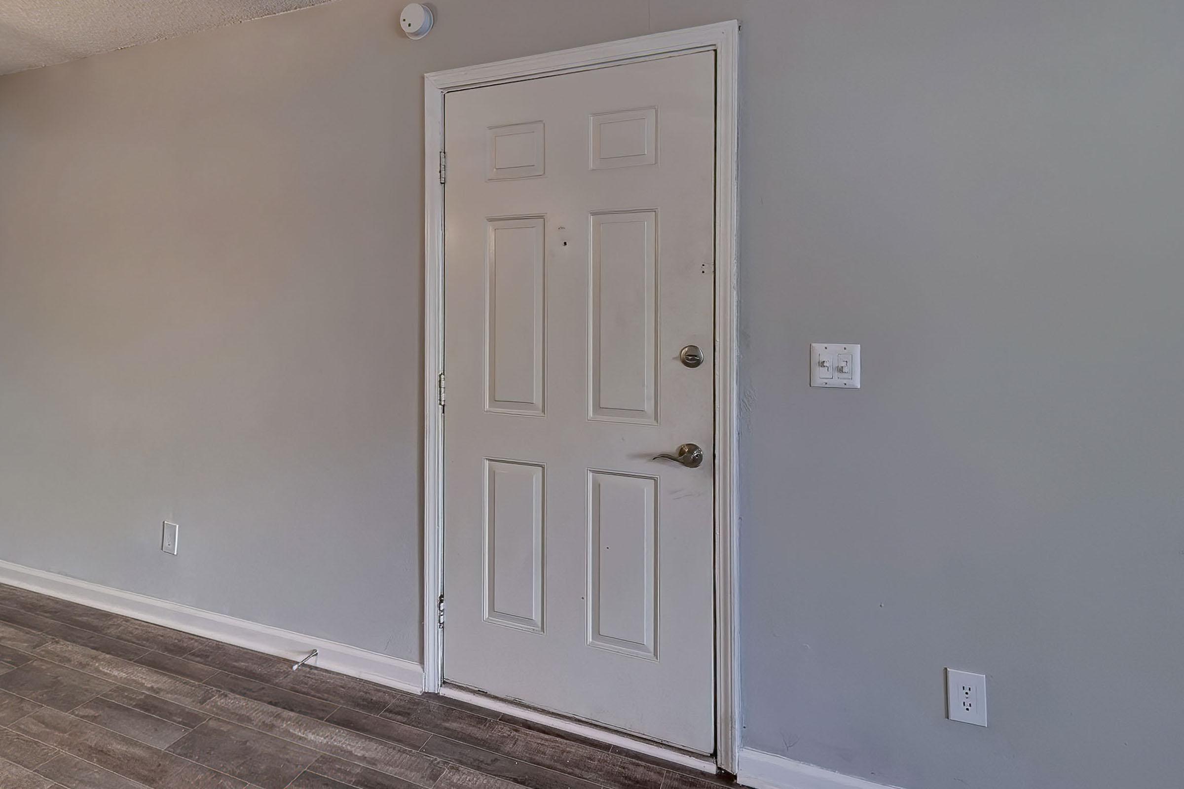 Interior view of a room featuring a white door against a light gray wall. The door has a round doorknob and a deadbolt lock. To the right, there is a light switch and an electrical outlet. The floor is covered with dark wood-like planks, providing a modern look to the space. Natural light illuminates the area.