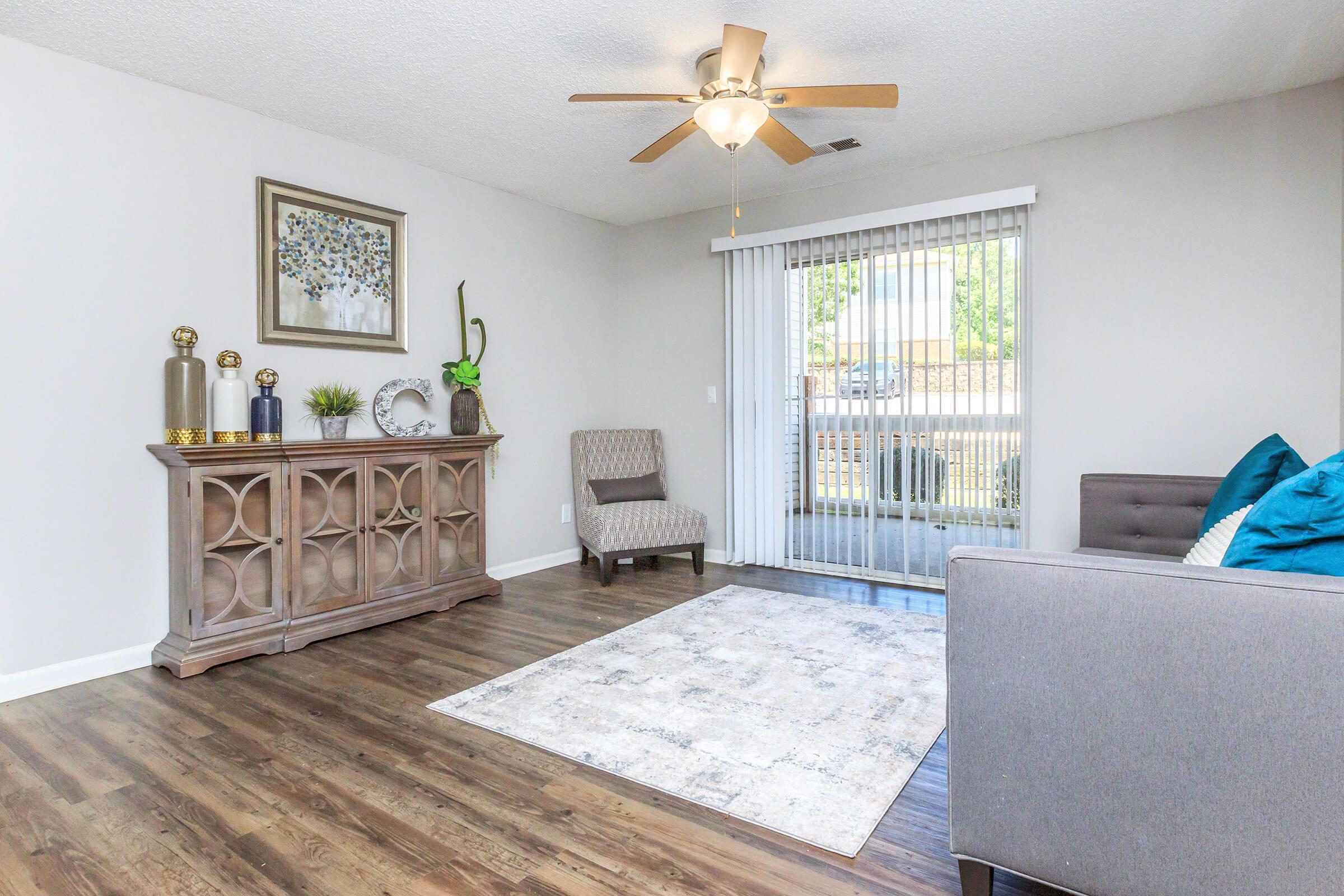 A modern living room featuring a gray sofa with blue accents, a neutral area rug, and a decorative wooden sideboard. A ceiling fan is mounted above, and large sliding glass doors allow natural light to enter, revealing an outdoor view. A cozy armchair and decorative plants complete the space.