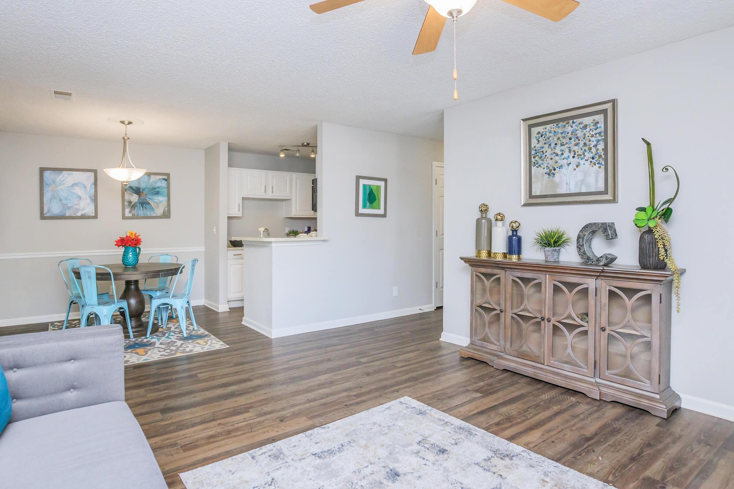 A spacious, modern living area featuring a comfortable gray sofa, a decorative area rug, and a wooden sideboard with various decorative items. There is a dining area with a round table and blue chairs, adjacent to a kitchen visible in the background. The walls are painted light gray, creating a bright and inviting atmosphere.