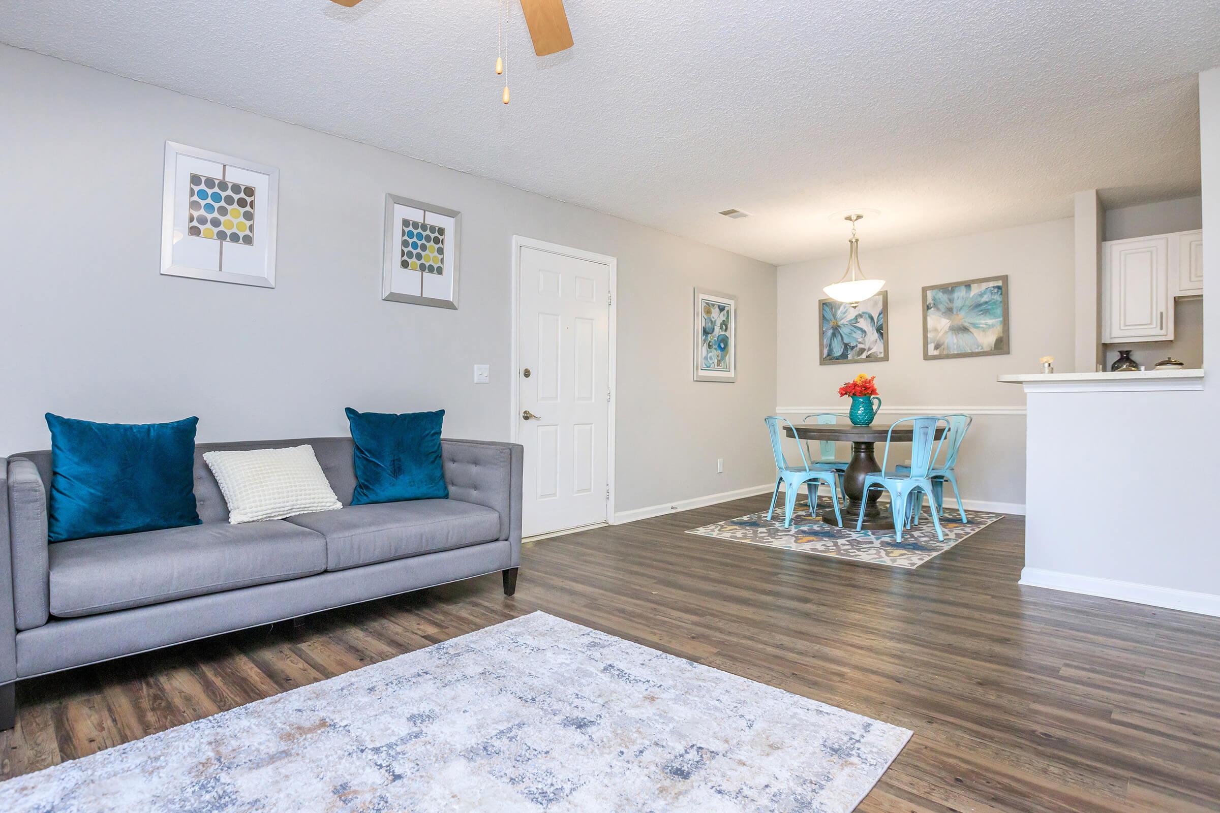 Modern living room featuring a gray sofa with blue accent pillows, a light-colored rug, and framed art on the walls. Adjacent dining area with a round table and blue chairs, illuminated by a pendant light. Neutral walls and hardwood floors create a bright, inviting atmosphere.