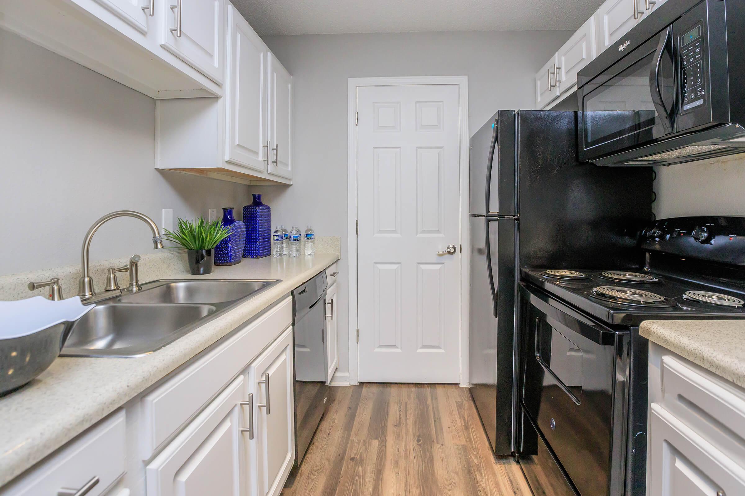 Modern kitchen with light countertops, white cabinetry, and black appliances, including an oven, microwave, and refrigerator. A double sink is on the left with a small plant and water bottles on the counter. A white door is visible at the end of the kitchen. The floor features wood-like flooring.