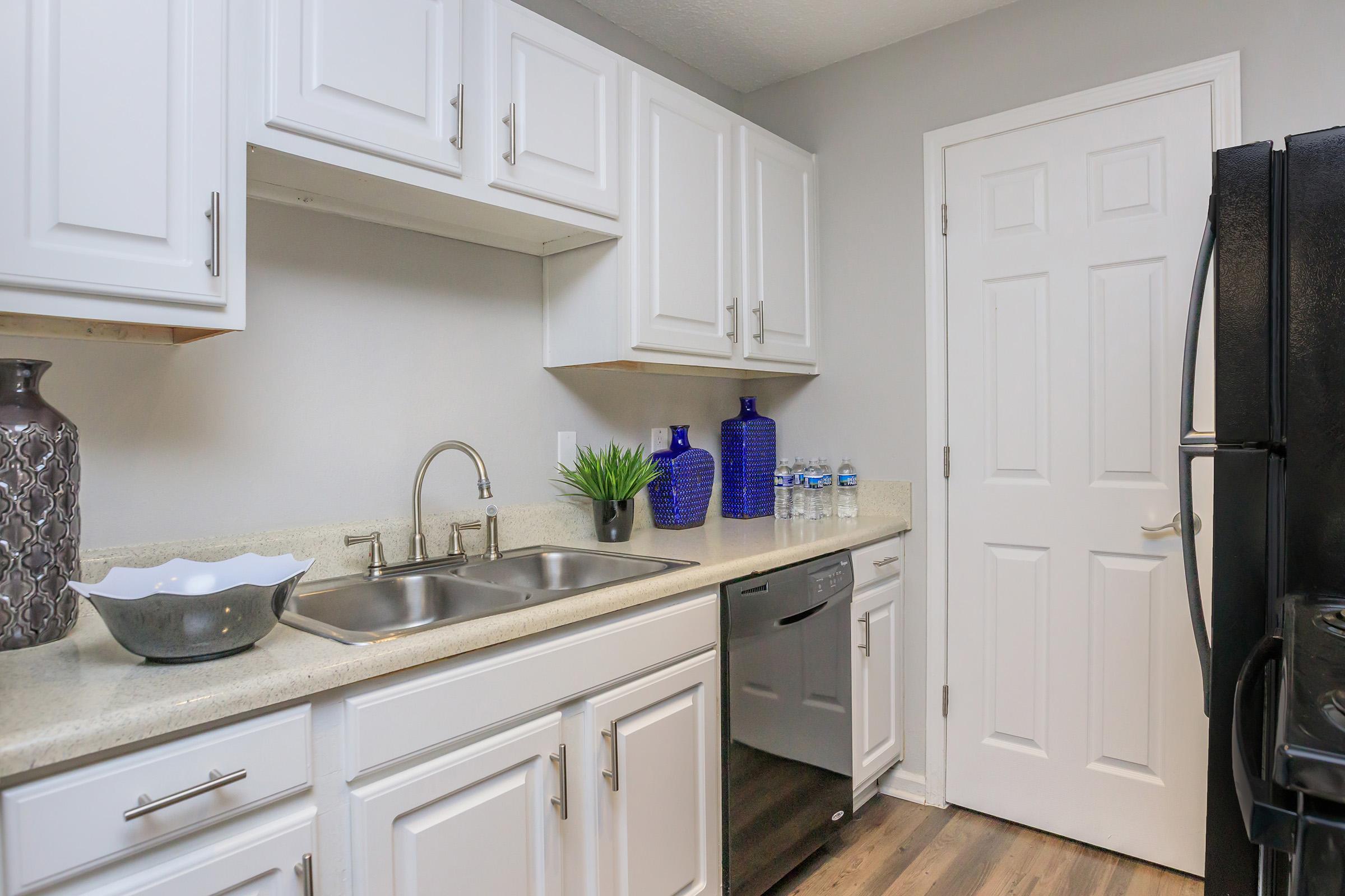 A modern kitchen featuring white cabinetry, a double sink, and a countertop with decorative items. There are blue vases and bottles on the counter, and stainless steel appliances, including a dishwasher and a black refrigerator. The walls are painted gray, creating a sleek and contemporary look.