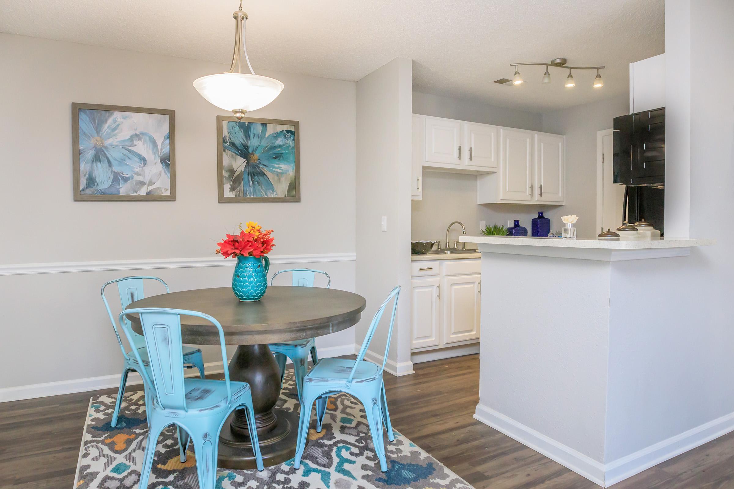 A modern dining area featuring a round table with a dark wood finish, surrounded by four light blue metal chairs. A turquoise vase with red flowers sits on the table. The room has gray walls with floral artwork and a partially visible kitchen area with white cabinets. A patterned rug adds color to the floor.