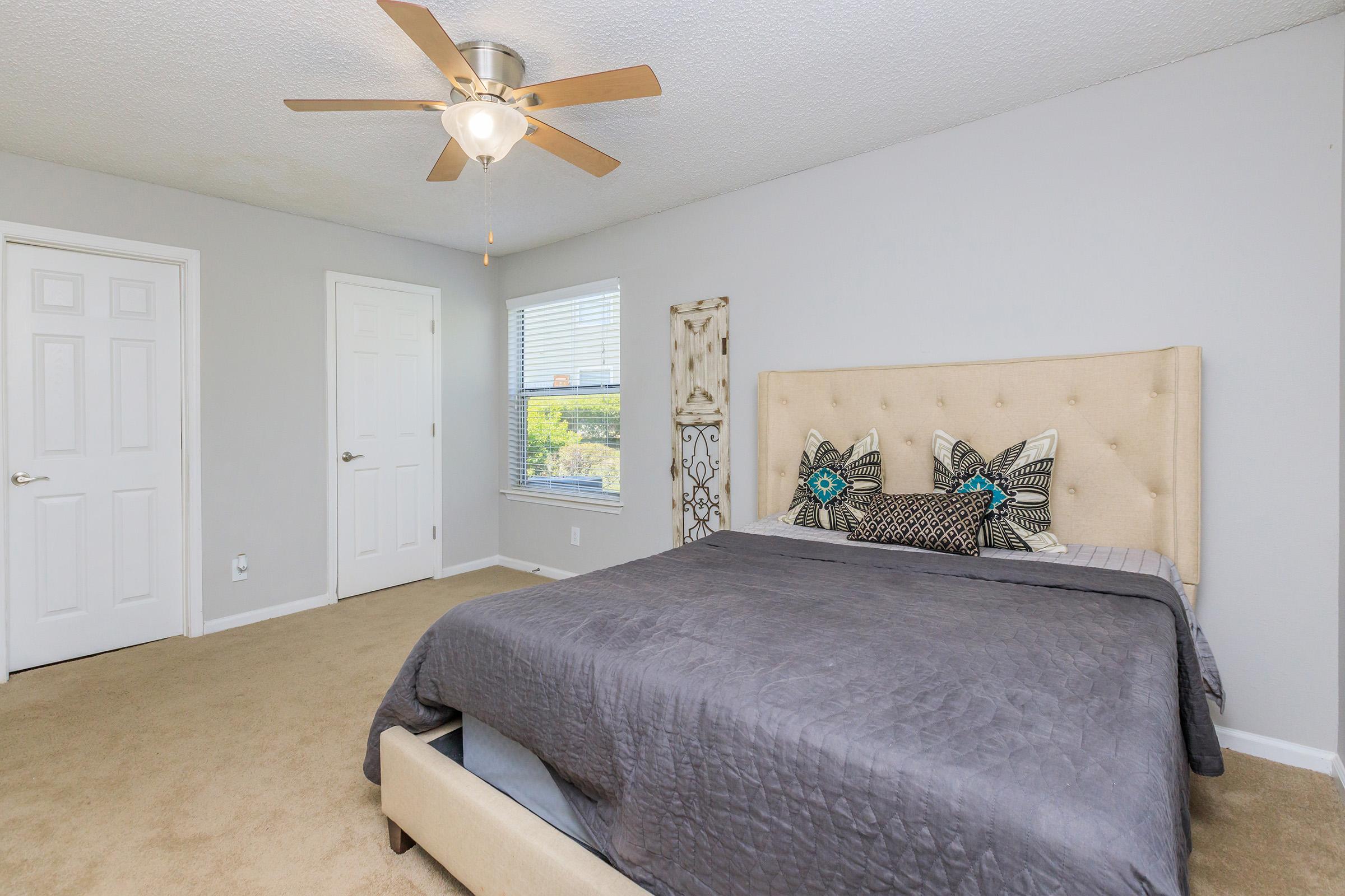 A cozy bedroom featuring a queen-sized bed with gray bedding and decorative pillows, a ceiling fan, beige walls, and two doors. A window with blinds allows natural light to illuminate the room, enhancing the inviting atmosphere.