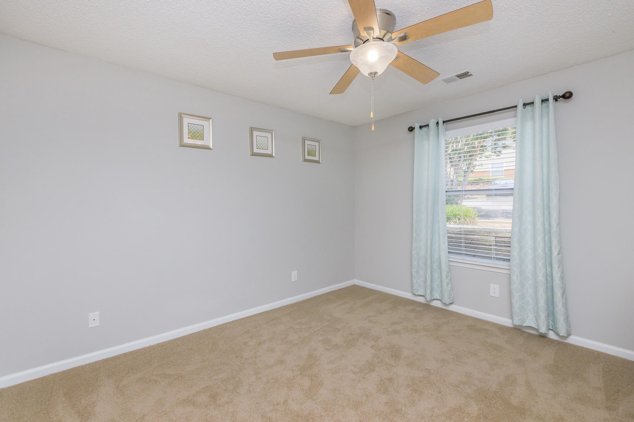 A clean and simple bedroom with light gray walls and beige carpet. A ceiling fan with wooden blades is installed. Three framed pictures are arranged on the wall above a window with light blue curtains, allowing natural light to enter the room. The space is well-lit and appears inviting and spacious.