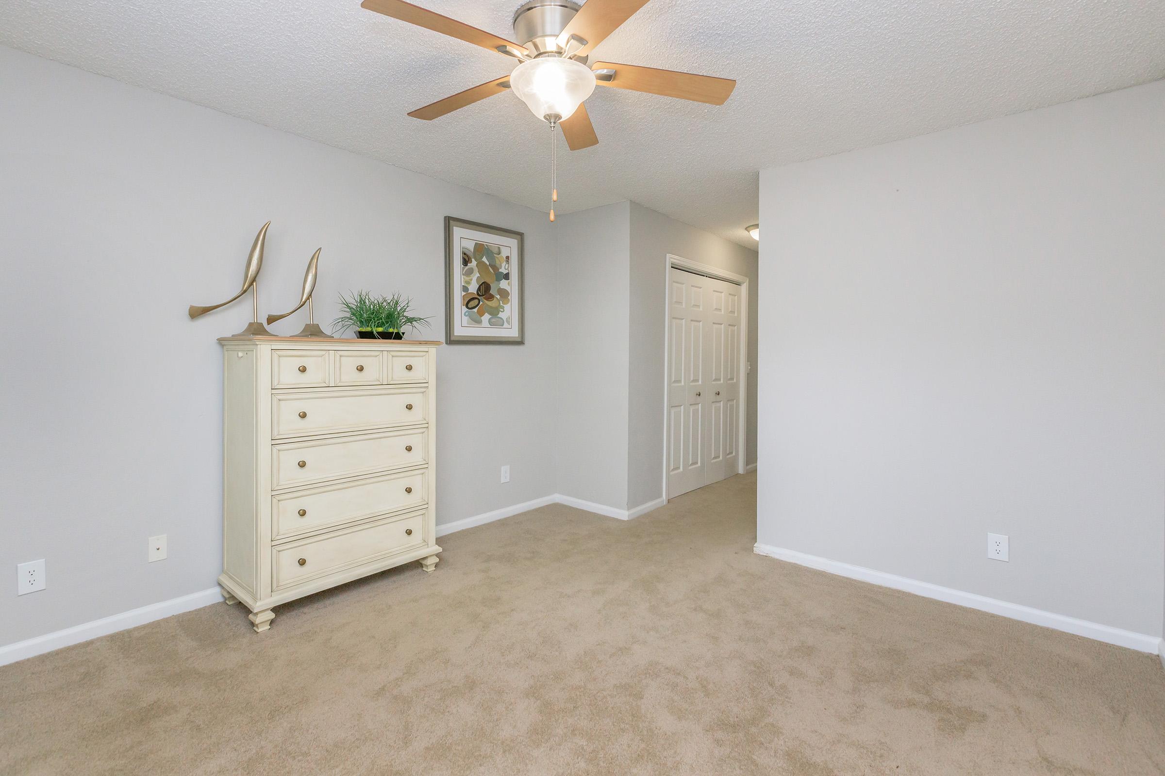 A well-lit room featuring light beige carpet, a ceiling fan with wooden blades, and a cream-colored dresser with six drawers. A framed artwork is displayed on the wall above the dresser, and a hallway with white doors can be seen in the background. The walls are painted a soft gray.