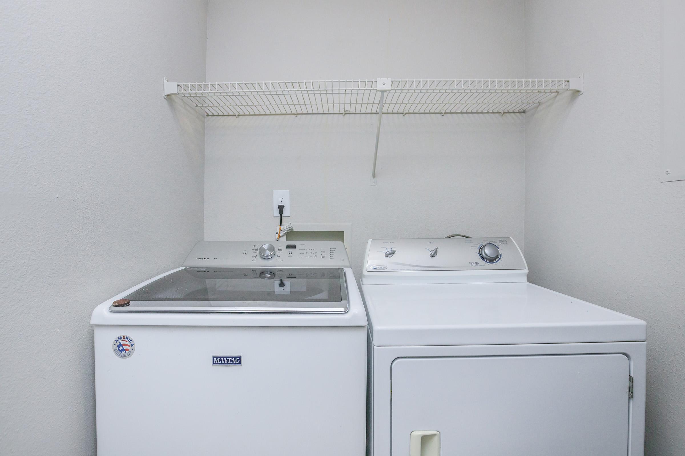 A laundry area featuring a washing machine and a dryer side by side, with a wire shelf above them. The walls are painted light, creating a clean and modern appearance.