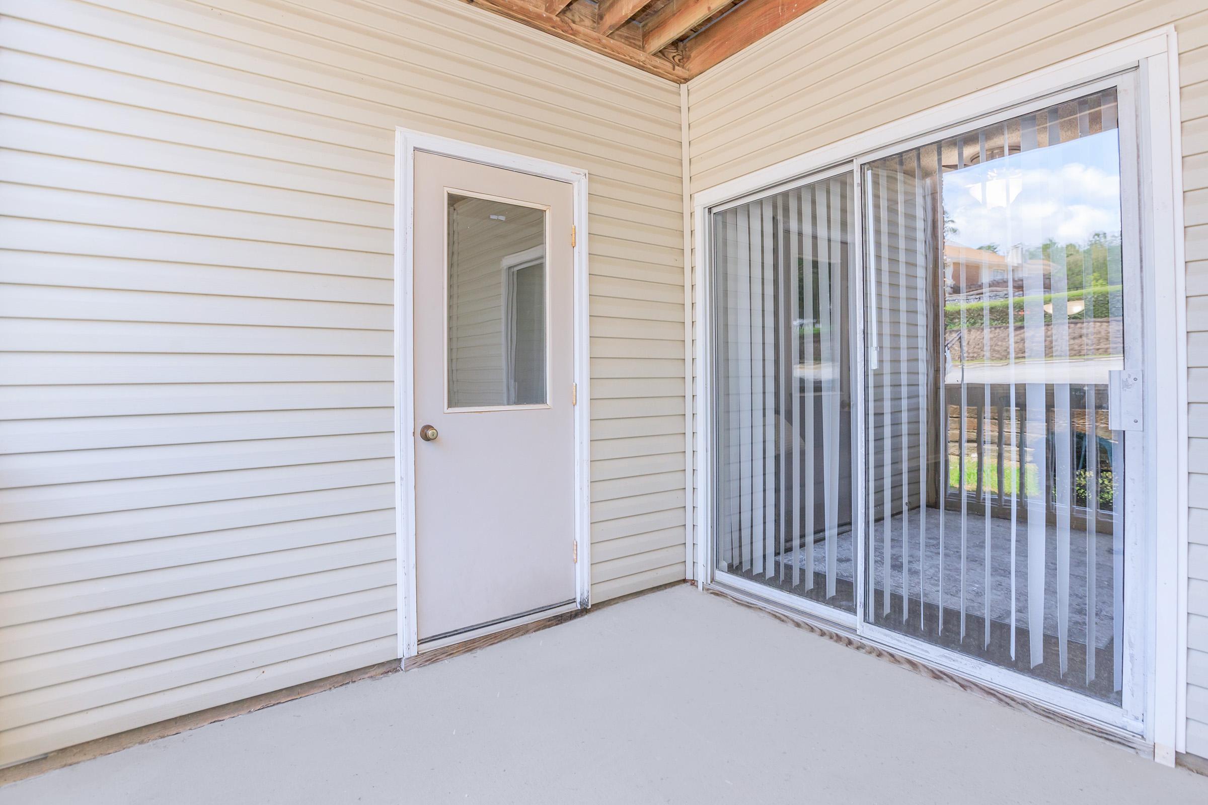 A wooden porch area featuring a door on the left and a large sliding glass door on the right with vertical blinds. The walls are lined with light-colored siding, and the floor is a smooth, light-colored concrete. The area is well-lit with natural light coming from outside.