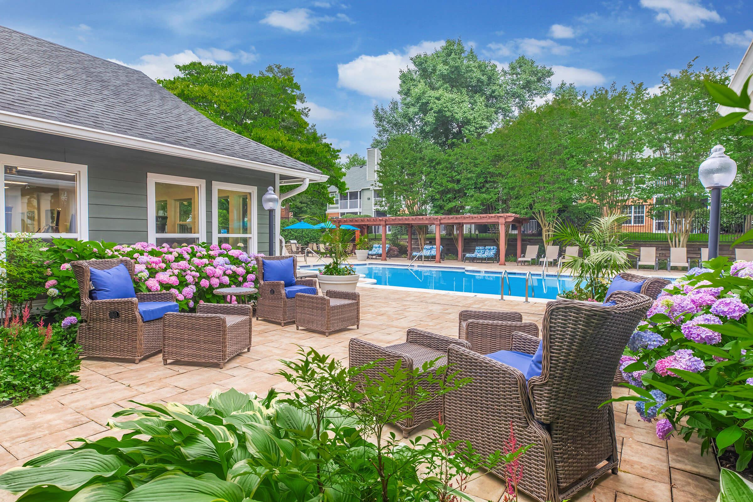 A beautifully landscaped outdoor area featuring a pool, surrounded by lush greenery and blooming flowers. Comfortable seating is arranged with blue cushions near the pool, creating a serene space for relaxation. The sky is clear, enhancing the inviting atmosphere of this garden setting.