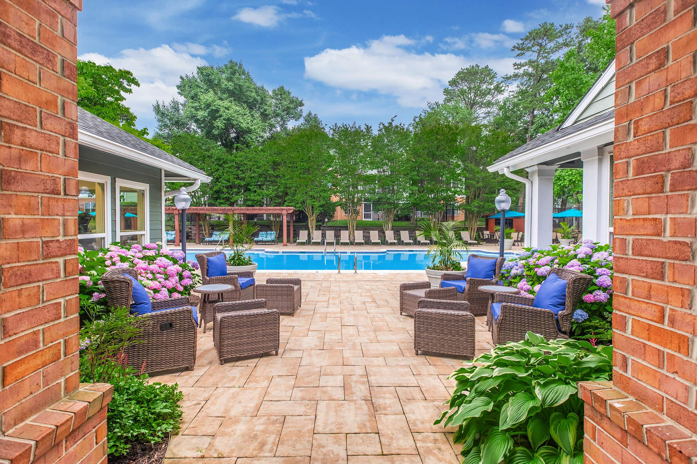 A serene pool area surrounded by lush greenery and flowering plants. Cozy wicker chairs with blue cushions are arranged on a stone patio, leading to a sparkling pool. In the background, there are lounge chairs and shade umbrellas, creating a relaxing outdoor atmosphere under a clear blue sky.