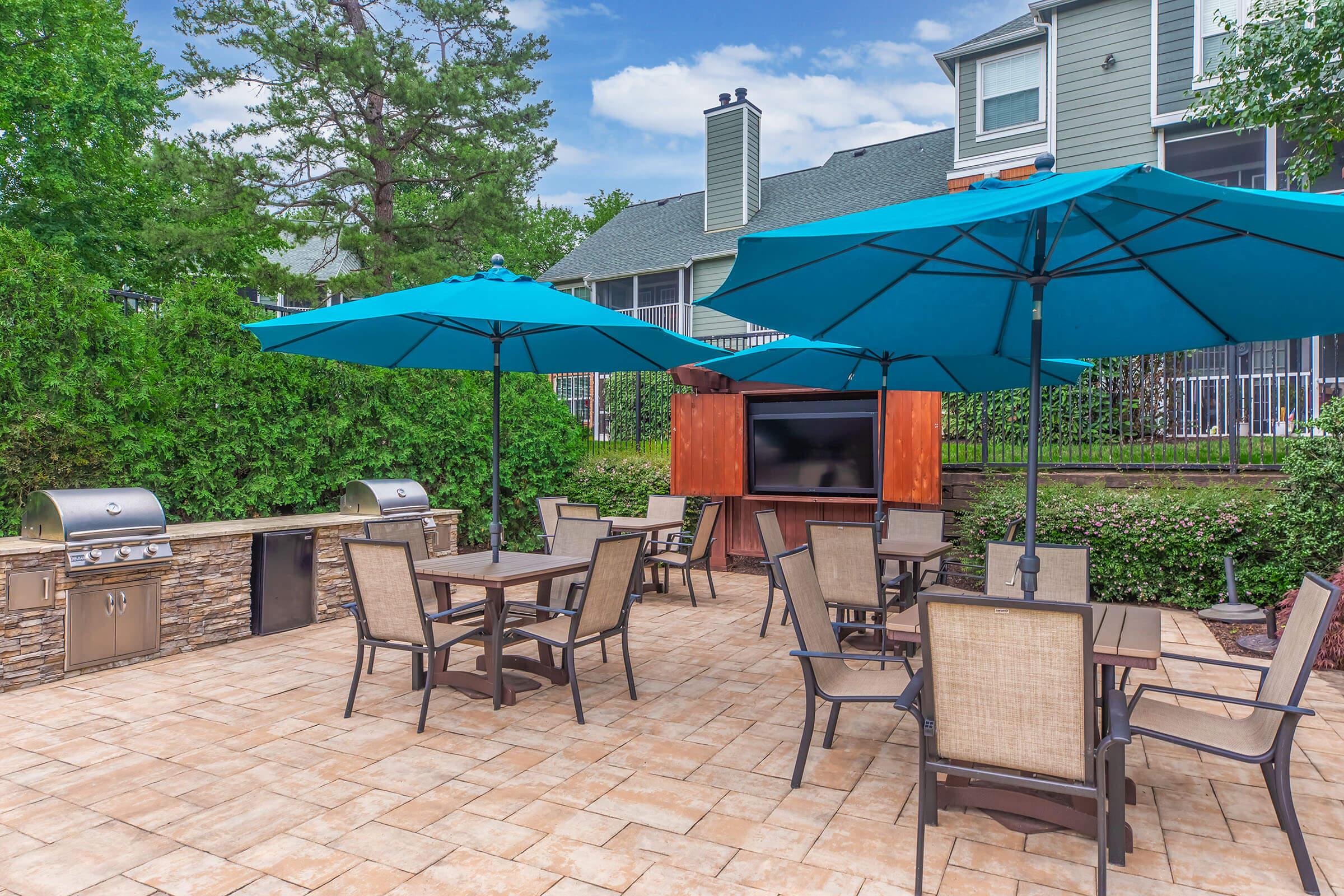 Outdoor patio area featuring a stone grill, two large blue umbrellas, and wooden tables and chairs. A flat-screen television is mounted on a nearby wooden structure, surrounded by green trees and shrubs. The background shows a building with multiple stories under a partly cloudy sky.