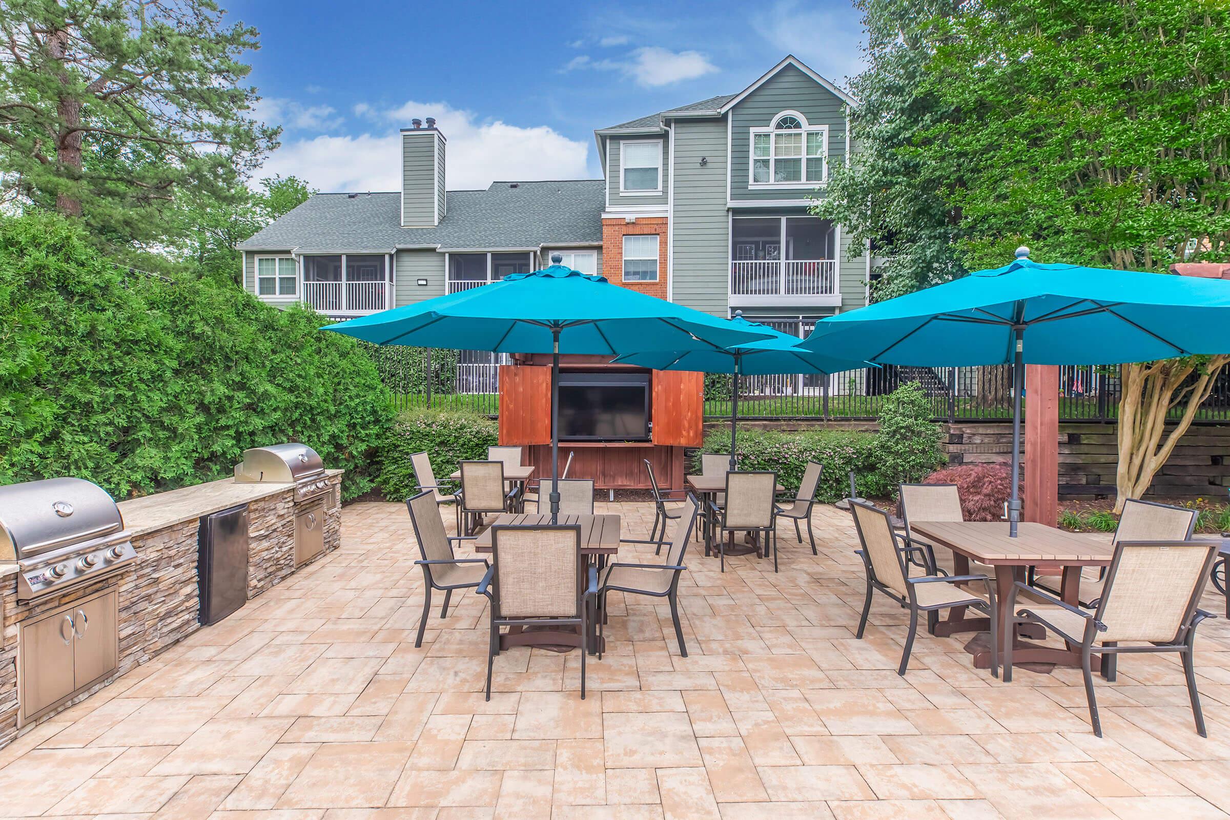 An outdoor patio area featuring several tables with chairs, large blue umbrellas for shade, a barbecue grill, and a mounted TV. In the background, there are residential buildings surrounded by greenery under a clear blue sky. The setting is inviting for social gatherings or relaxation.