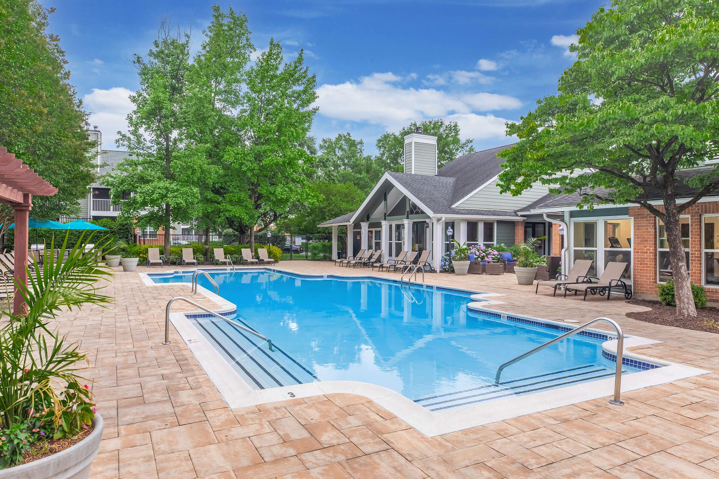 Outdoor swimming pool surrounded by lounge chairs, with a backdrop of a clubhouse and lush green trees. The pool features a shallow entry, and the area is well-maintained with decorative planters and an inviting atmosphere, perfect for relaxation.