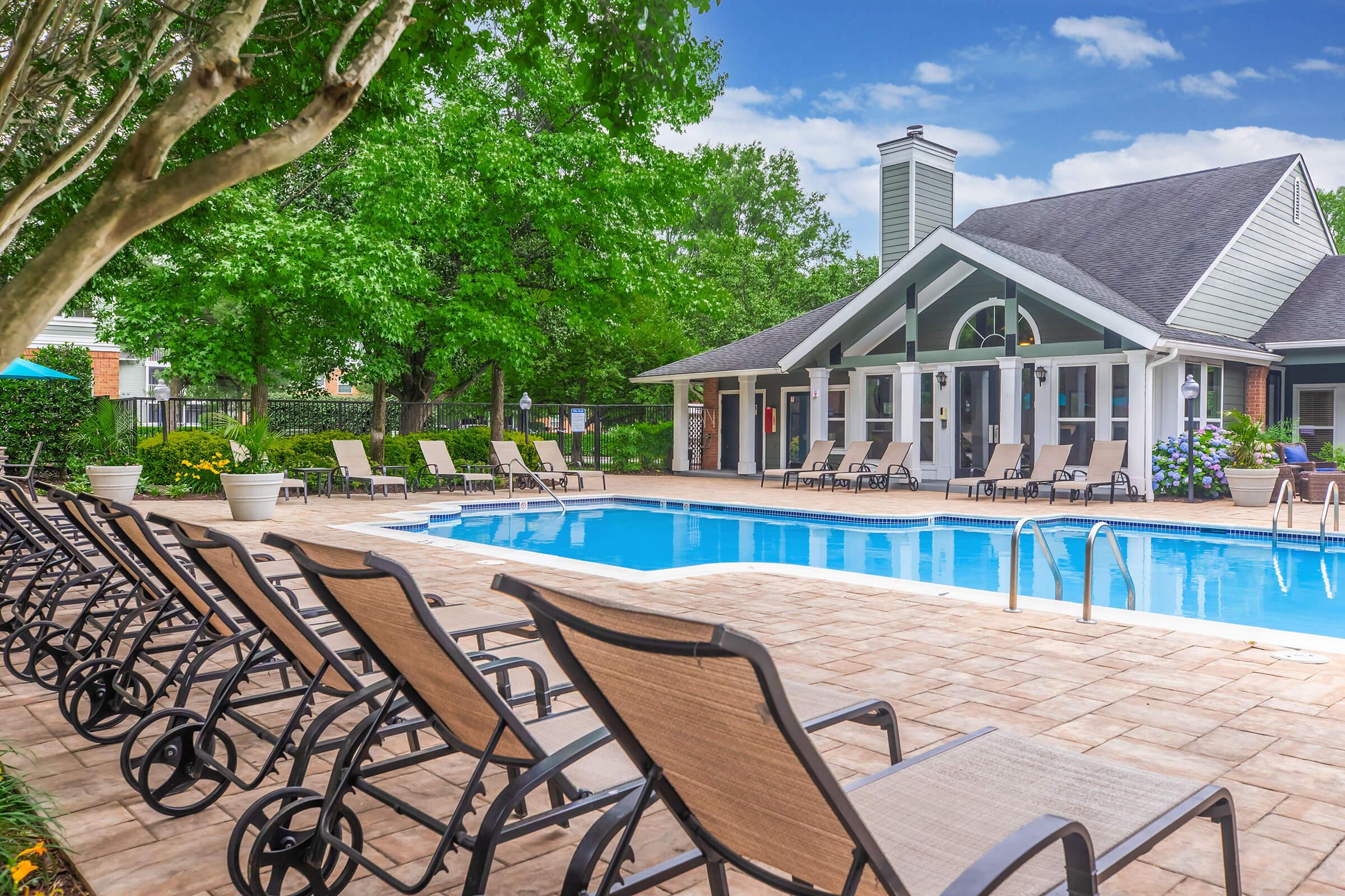 A sunny outdoor swimming pool area featuring lounge chairs lined up on a patio. In the background, there is a modern building with large windows, surrounded by lush greenery and trees. The sky is clear with a few clouds, creating a pleasant atmosphere for relaxation and leisure.