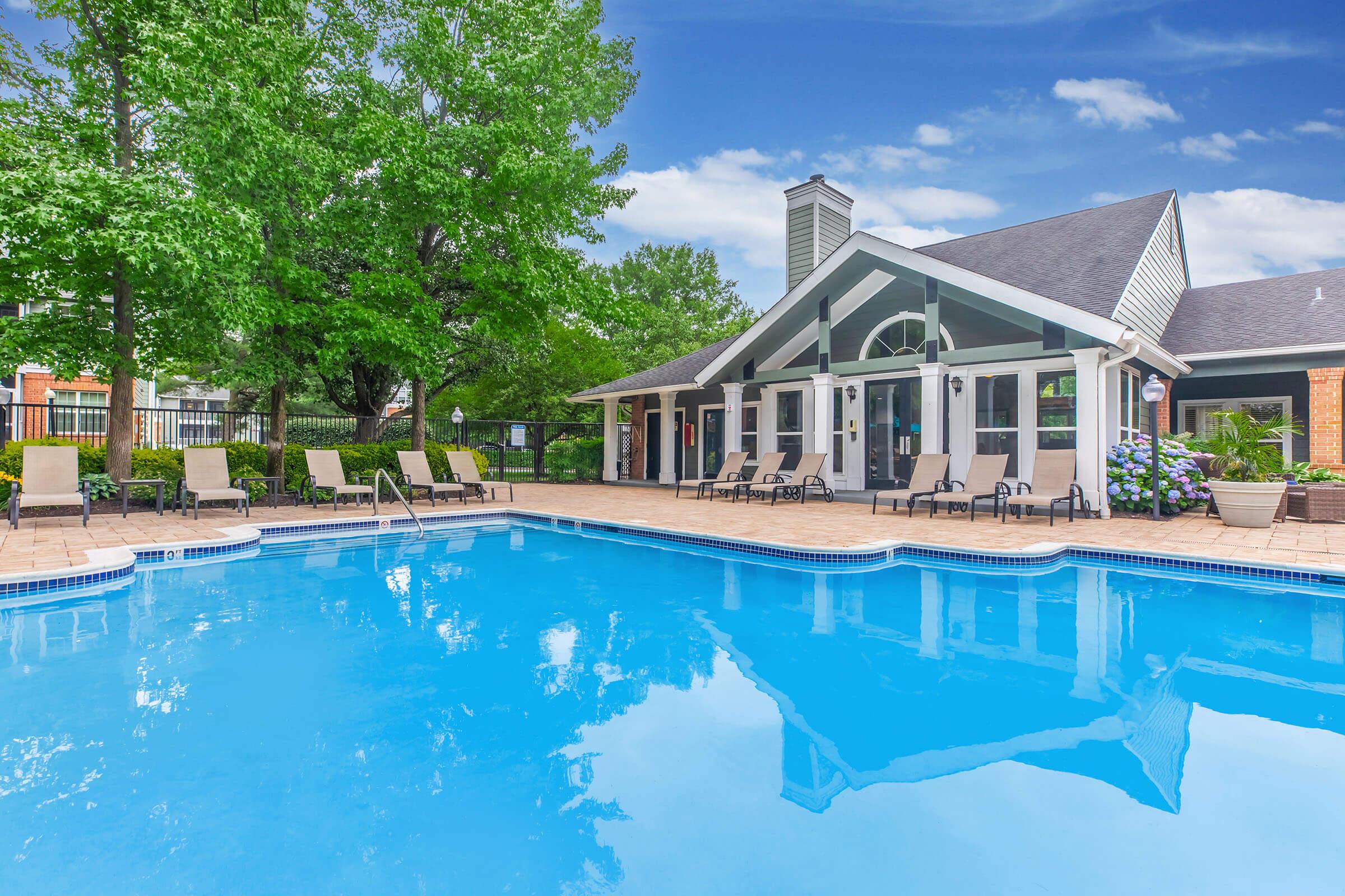 A clear blue swimming pool surrounded by lounge chairs and green trees. In the background, there is a modern clubhouse with large windows and a sloped roof. The sky is bright with a few clouds, creating a serene and inviting atmosphere.