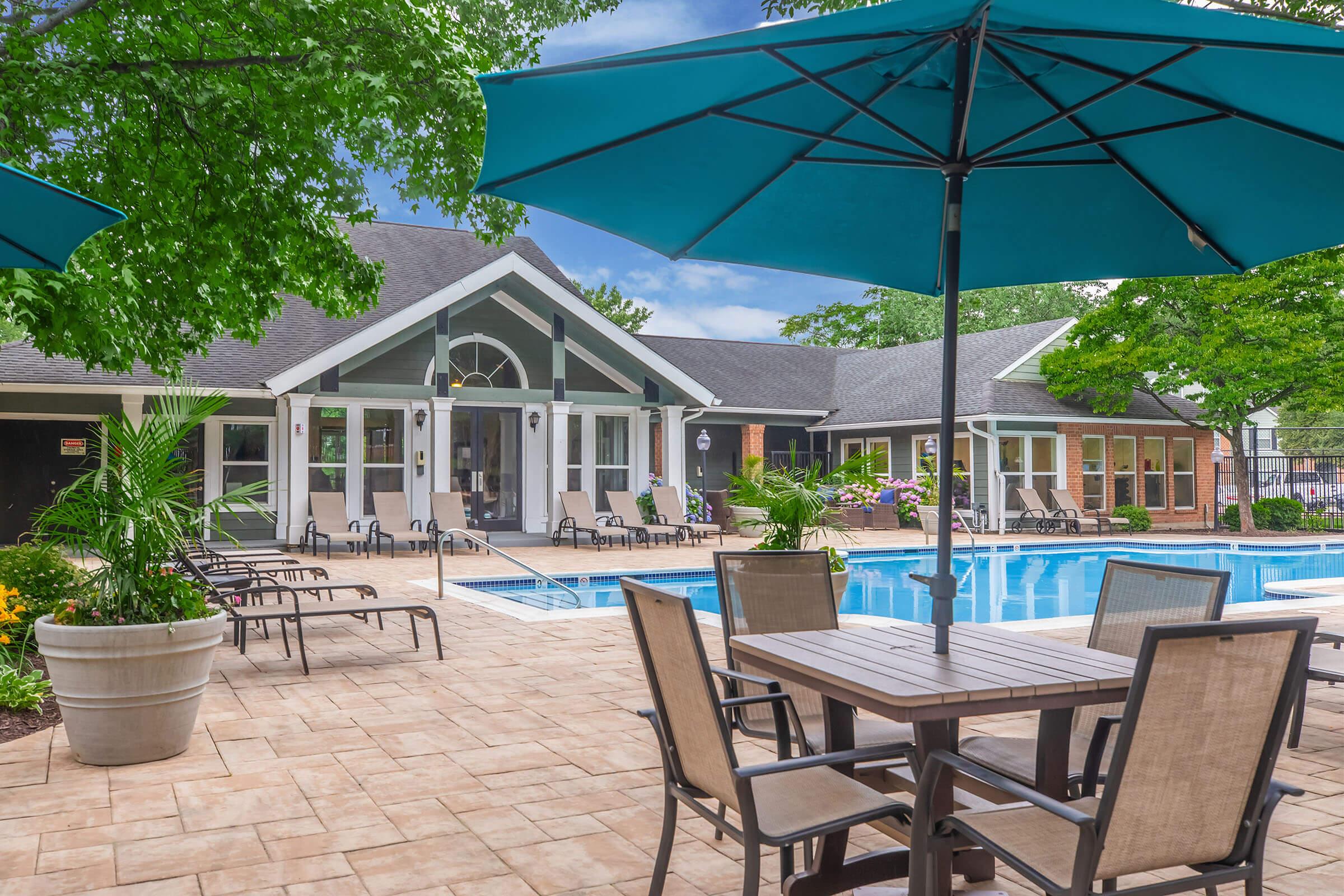 A sunny pool area featuring turquoise umbrellas over wooden tables and chairs. Lush green trees and colorful flowers surround the space. In the background, a modern building with large windows and a patio is visible, creating a welcoming atmosphere for relaxation and leisure by the pool.
