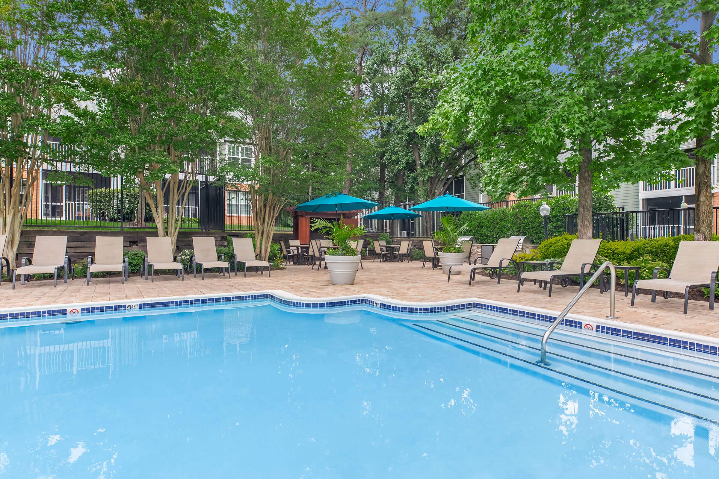 A tranquil pool area surrounded by lush greenery, featuring loungers and blue umbrellas. The clear blue water is inviting, and well-maintained landscaping enhances the relaxing atmosphere. Ideal for leisure and outdoor enjoyment.