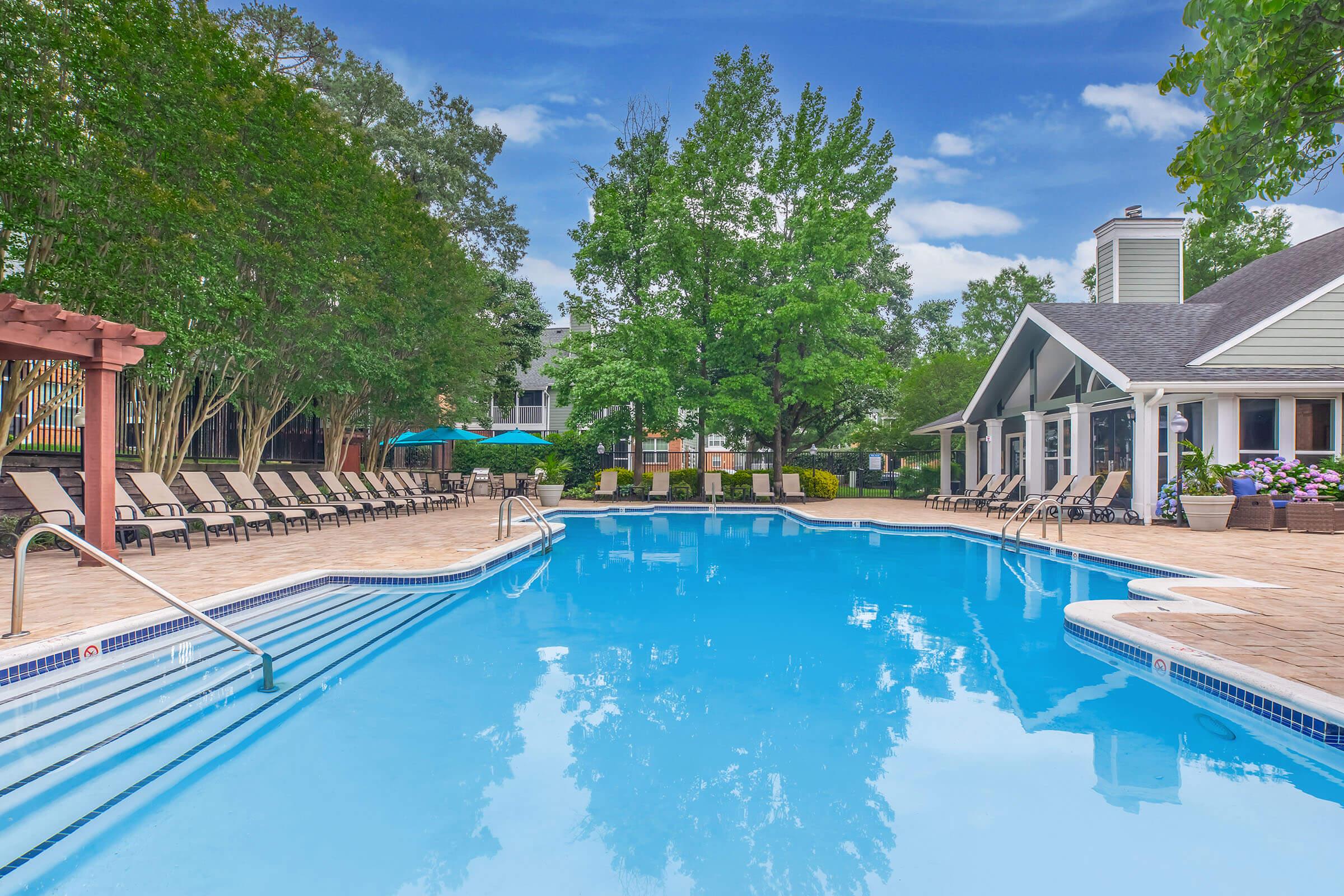 A serene swimming pool surrounded by lounge chairs and lush greenery. The water is clear and reflects the blue sky with clouds. There are shaded seating areas and a building in the background, creating a relaxing atmosphere ideal for leisure and recreation.