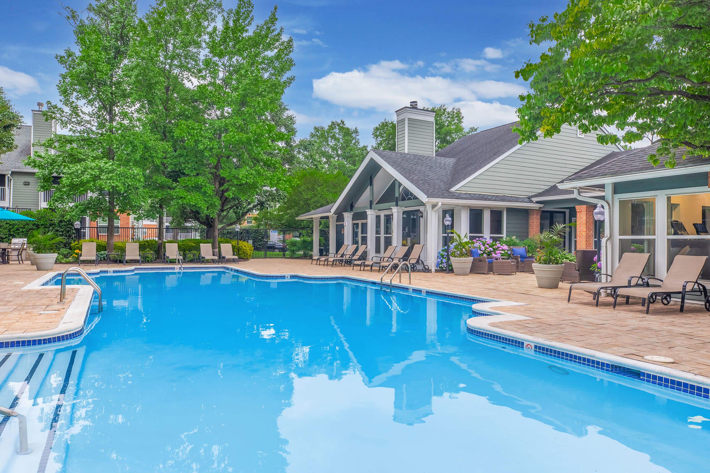 A clear blue swimming pool surrounded by lounge chairs and lush greenery. The pool area features a spacious deck, with trees and well-maintained landscaping in the background. A building with large windows is visible, contributing to a serene and inviting atmosphere.