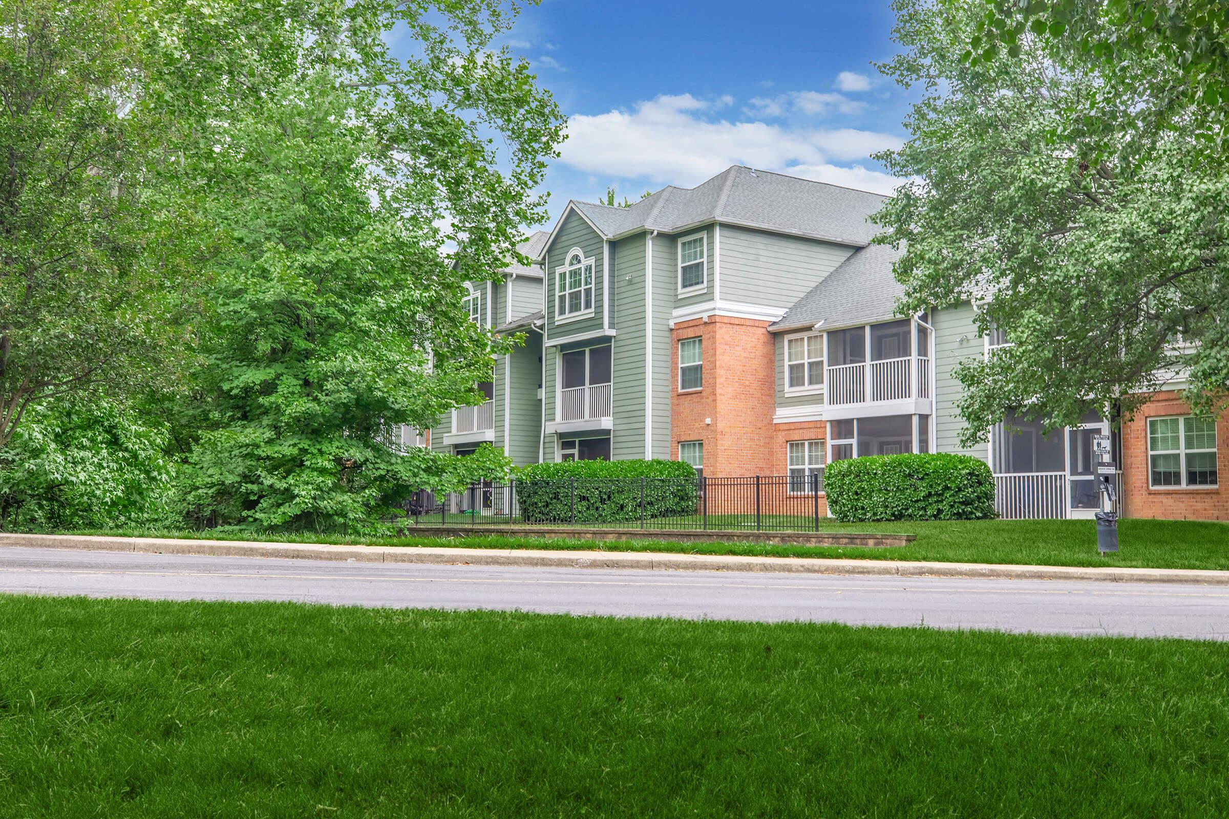 A modern multi-unit residential building with a mix of brick and siding, surrounded by lush green trees and grass. The building features large windows and balconies, set against a clear blue sky. A paved road runs in front of the property, enhancing its accessibility.