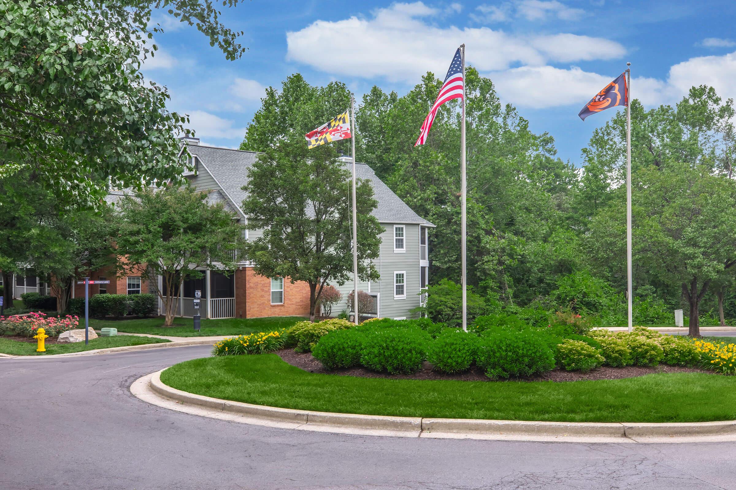 A scenic view of a landscaped area featuring a roundabout surrounded by greenery. In the background, there are two buildings with flagpoles displaying American and organizational flags. Lush trees and colorful flowerbeds enhance the natural beauty of the setting, creating a welcoming environment.