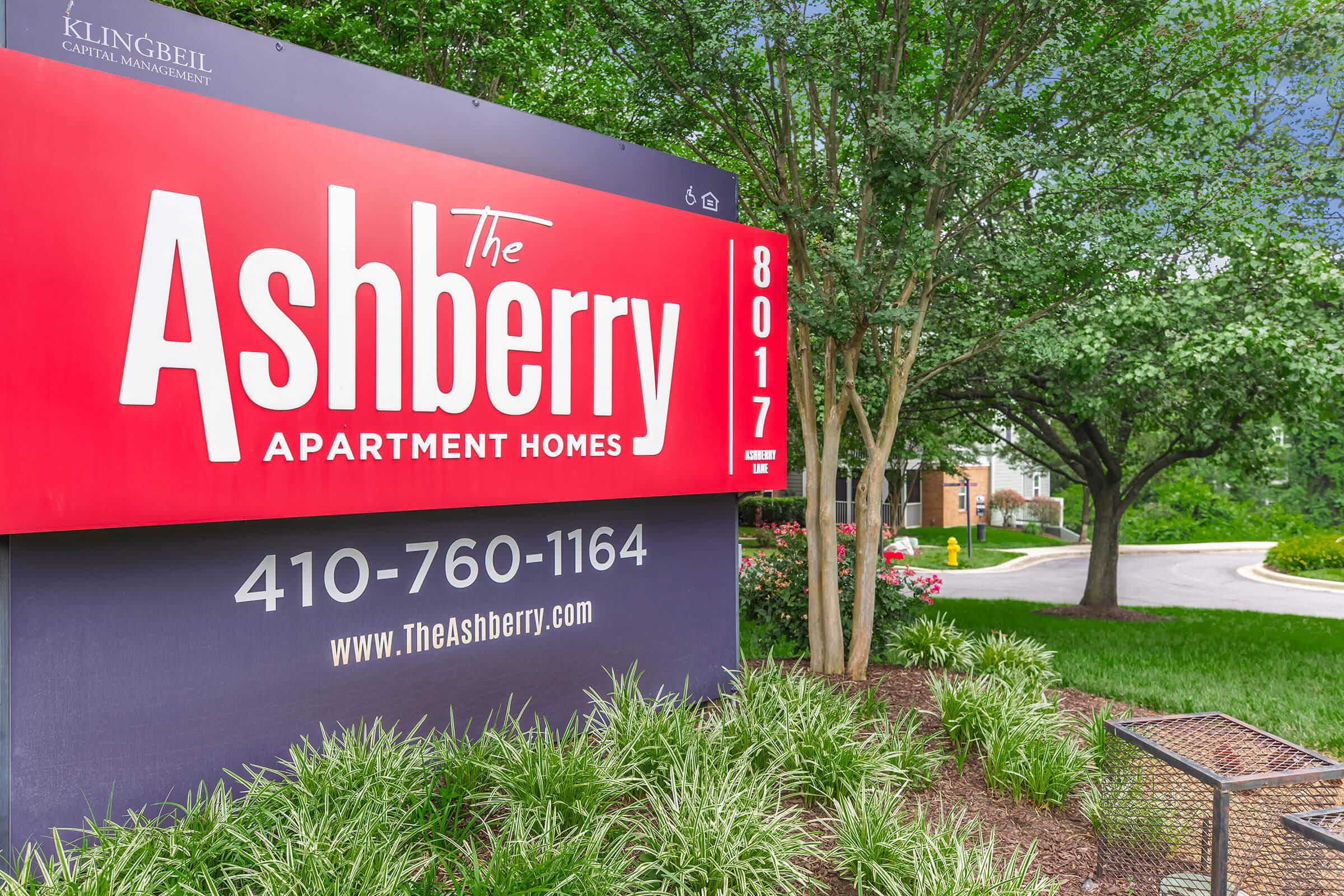 Sign for The Ashberry Apartment Homes displaying contact number 410-760-1164, located at 8017. The sign features a bold red background with white text. Surrounding the sign are trees and landscaped greenery, creating a welcoming entrance to the apartment complex.