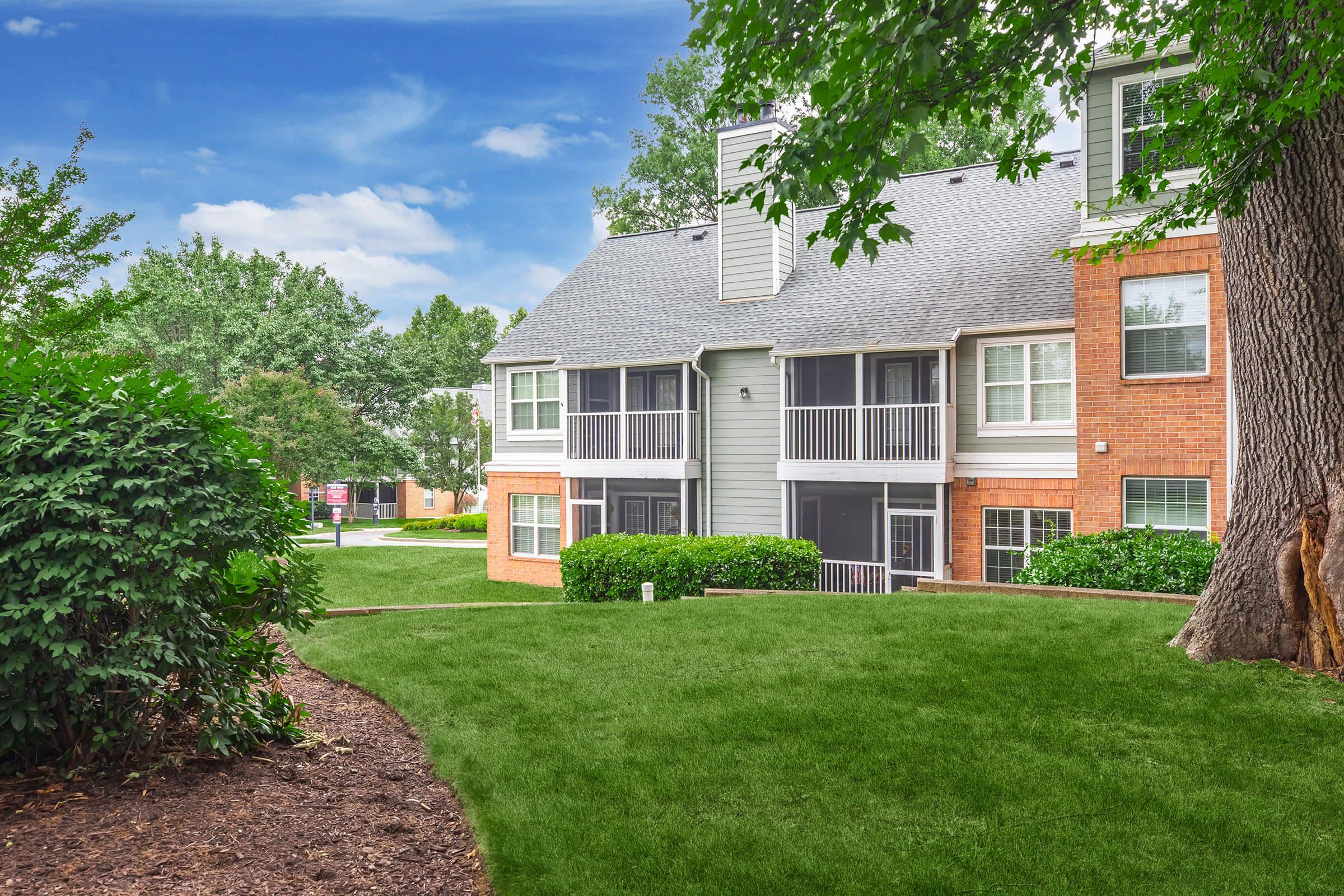 A landscaped area featuring a multi-unit residential building with gray siding and brick accents. The building has several balconies and large windows, surrounded by well-maintained greenery and grass. In the background, there are trees and other buildings, creating a peaceful neighborhood atmosphere.