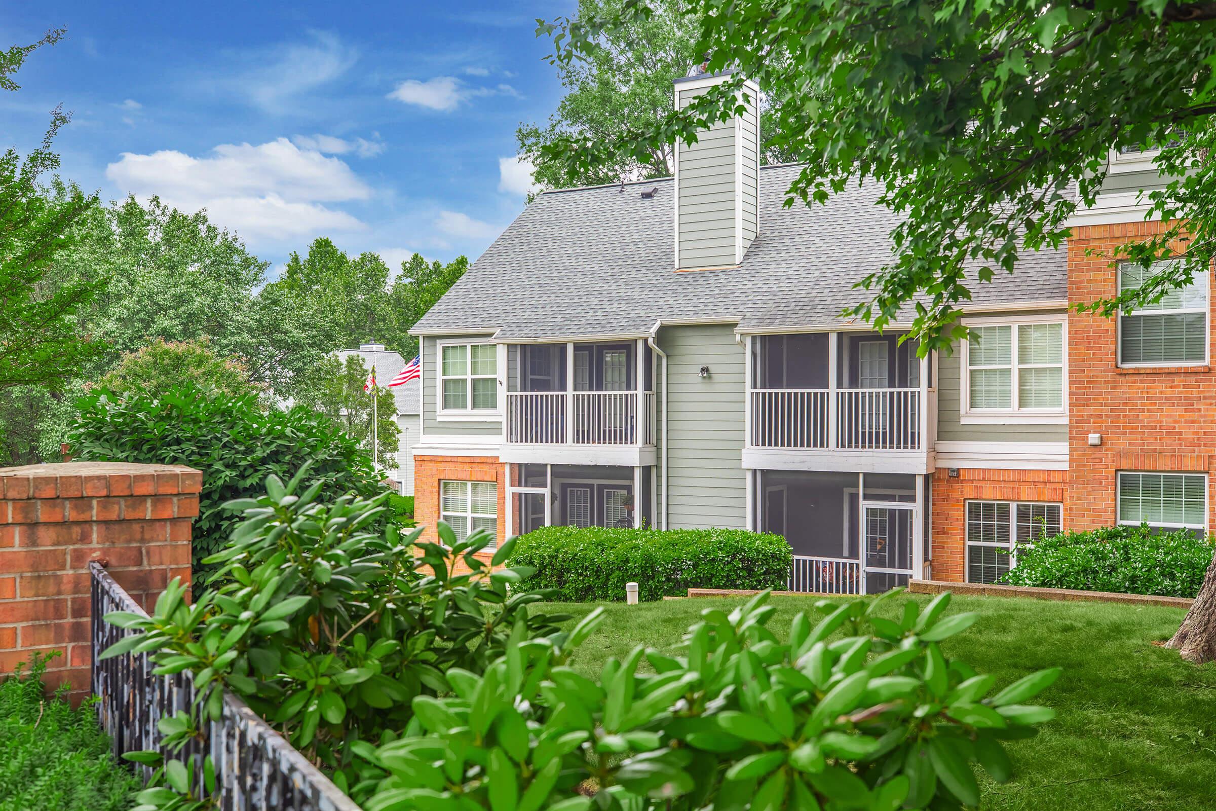 A well-maintained apartment building featuring a mix of brick and grey siding, with several balconies. Lush greenery surrounds the building, including bushes and trees. A flag can be seen in the background, and the sky is partially cloudy, adding a pleasant ambiance to the scene.