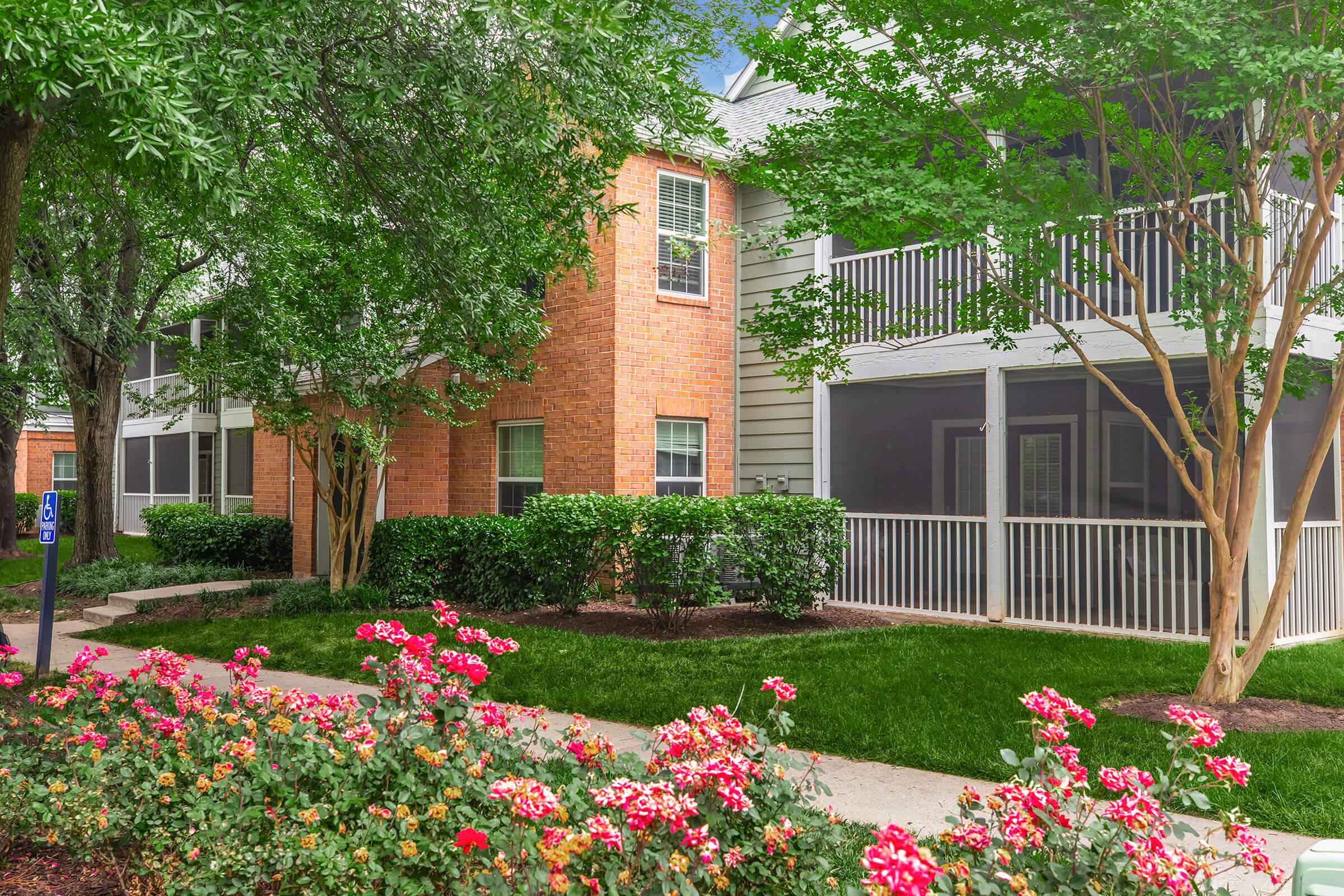 A landscaped exterior view of an apartment complex featuring brick and siding architecture. Lush greenery, including trees and bushes, surrounds the property, while vibrant flower beds with colorful blooms add charm. A screened-in porch is visible on the upper level, enhancing the inviting atmosphere.