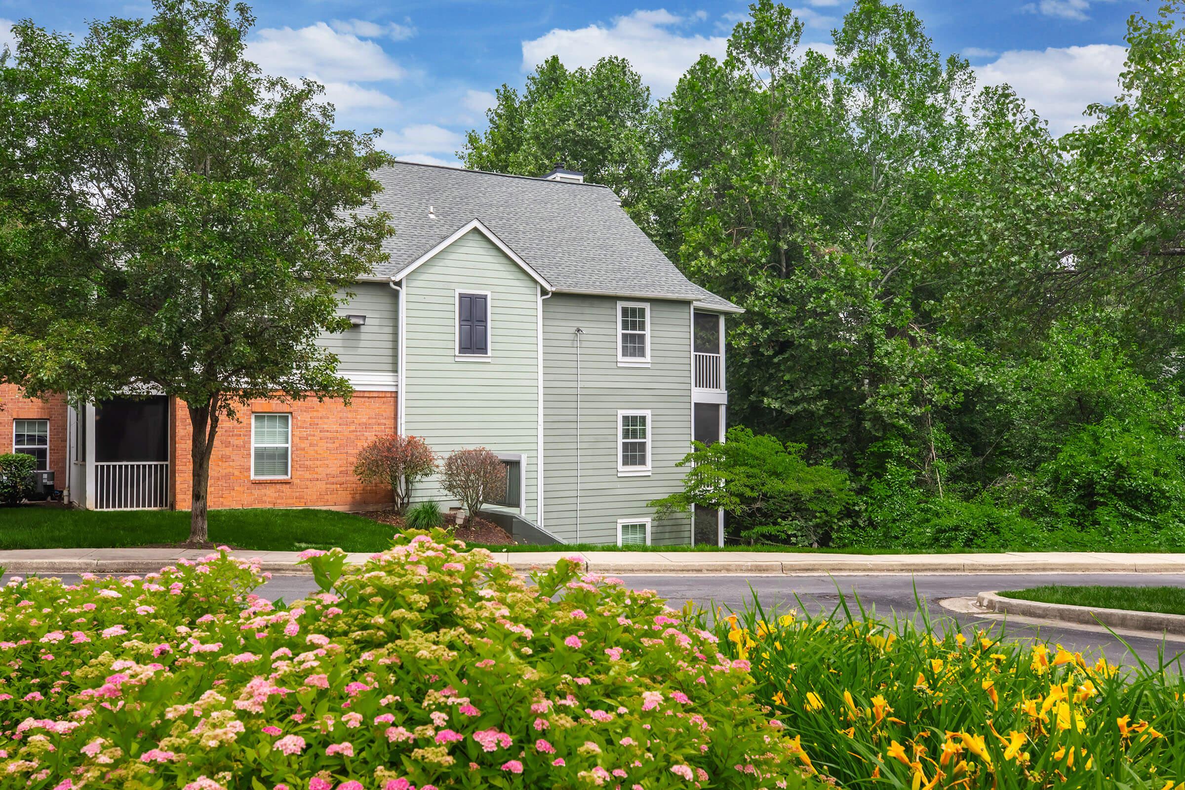 A peaceful residential building surrounded by lush greenery. In the foreground, colorful flowers bloom along a winding pathway. The architecture features a combination of brick and light green siding, with large windows that blend harmoniously with the natural landscape. Bright blue sky and trees provide a serene backdrop.