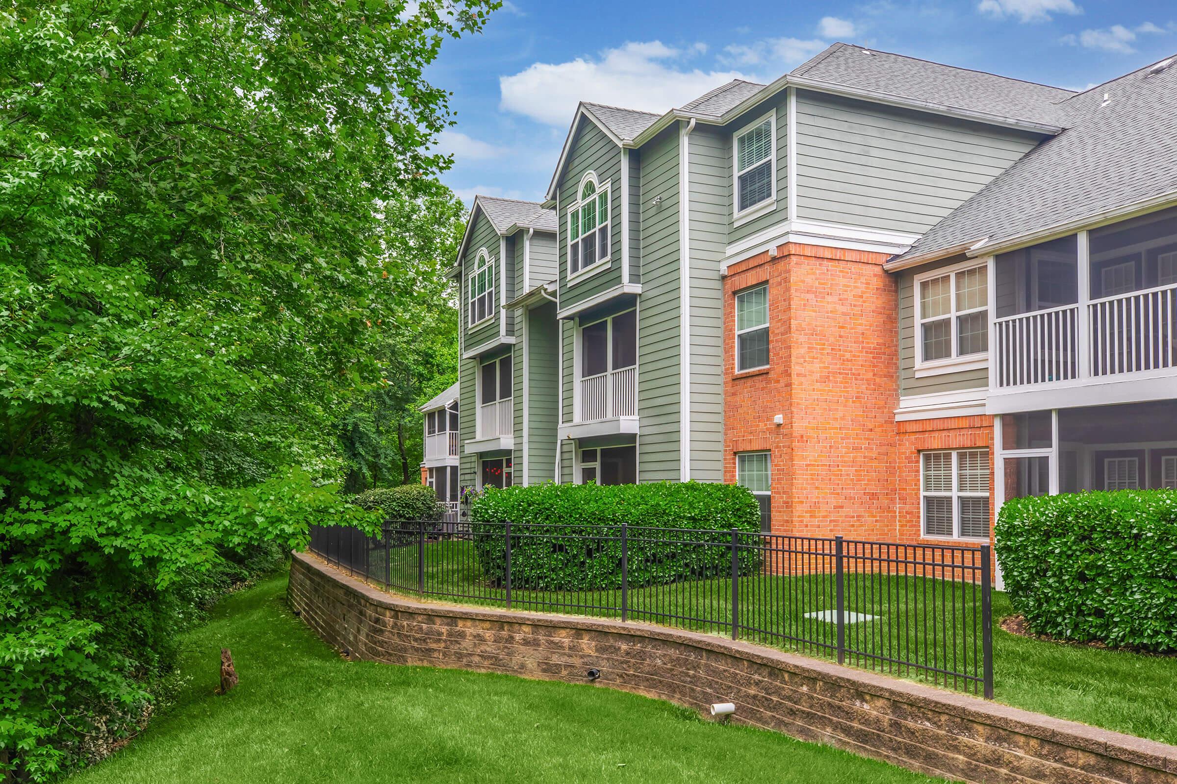 A modern apartment complex with a mix of green siding and brick exteriors, nestled among lush greenery. The buildings feature multiple levels, balconies, and a manicured lawn with a fenced perimeter. Bright blue skies are visible in the background.