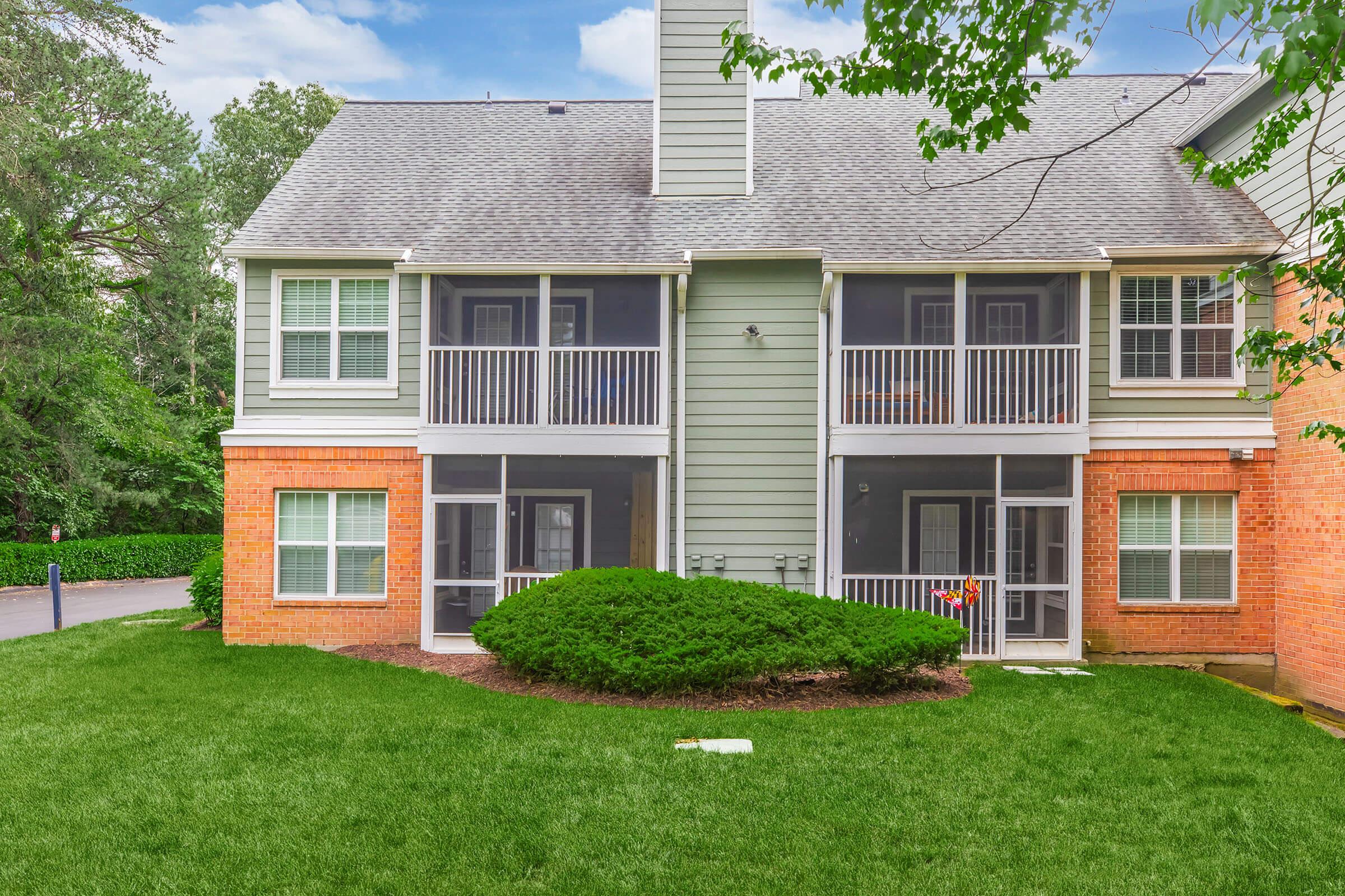Exterior view of a two-story apartment building featuring multiple balconies and large windows, surrounded by green grass and a neatly trimmed bush. The building has a mix of brick and siding materials, with a tree line in the background and a clear blue sky above.