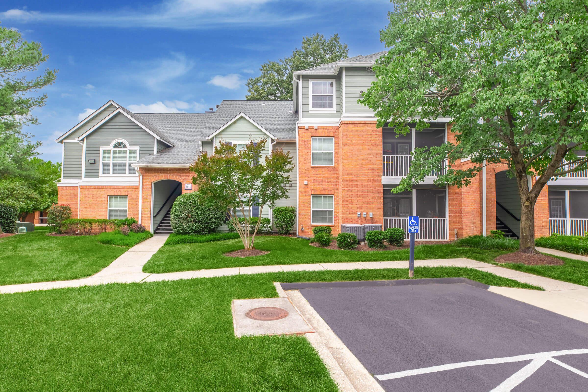 Modern apartment building with a mix of brick and siding exterior. The structure has multiple levels, featuring balconies, landscaped lawns, and a walkway leading to the entrance. Parking lot visible in front with accessible parking space. Surrounding greenery enhances the aesthetic appeal.