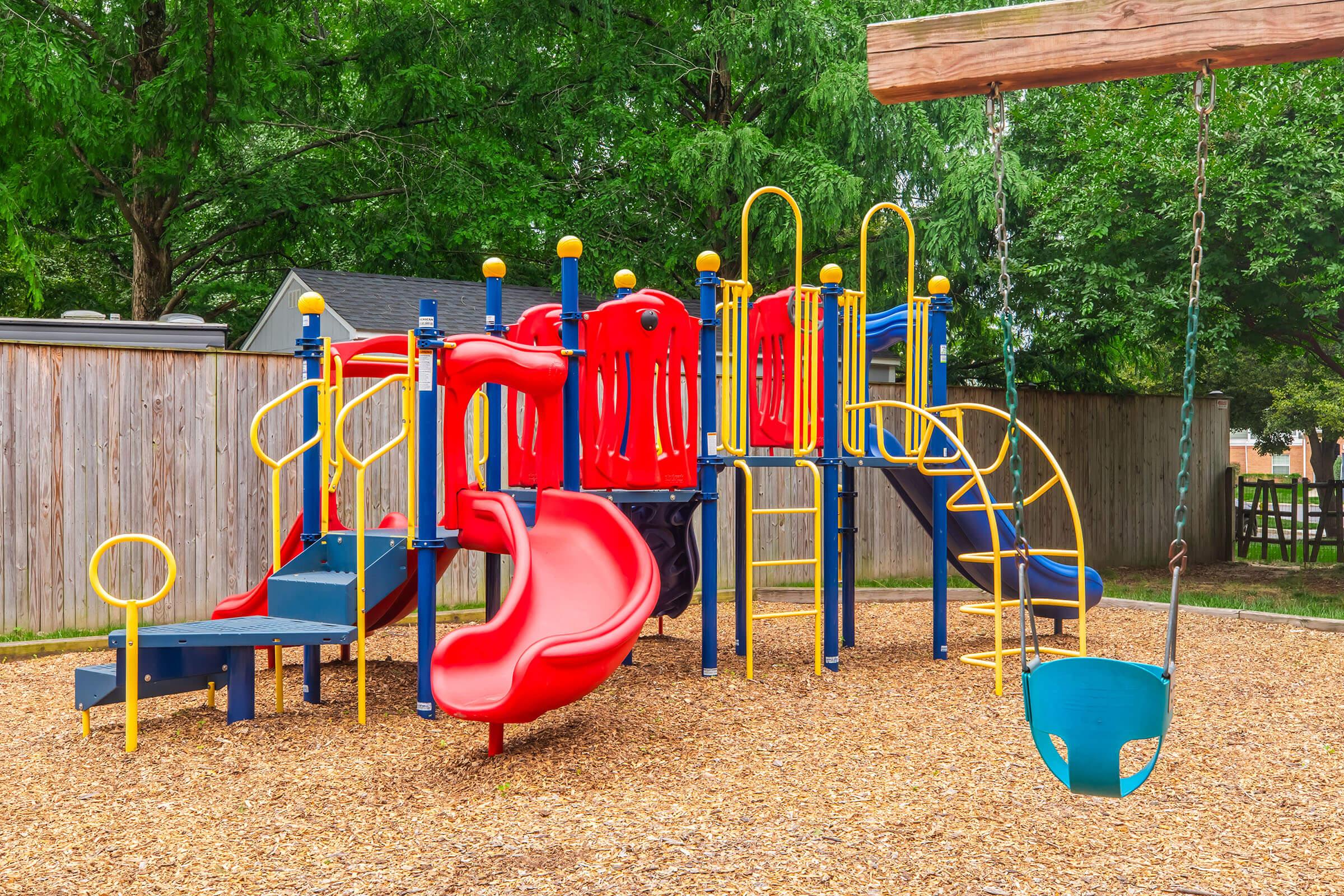A colorful playground featuring a climbing structure with slides, ladders, and swings, surrounded by mulch and trees. The play equipment is predominantly red and yellow, with blue swings, set in a safe, enclosed area. The scene conveys a fun and inviting atmosphere for children to play.