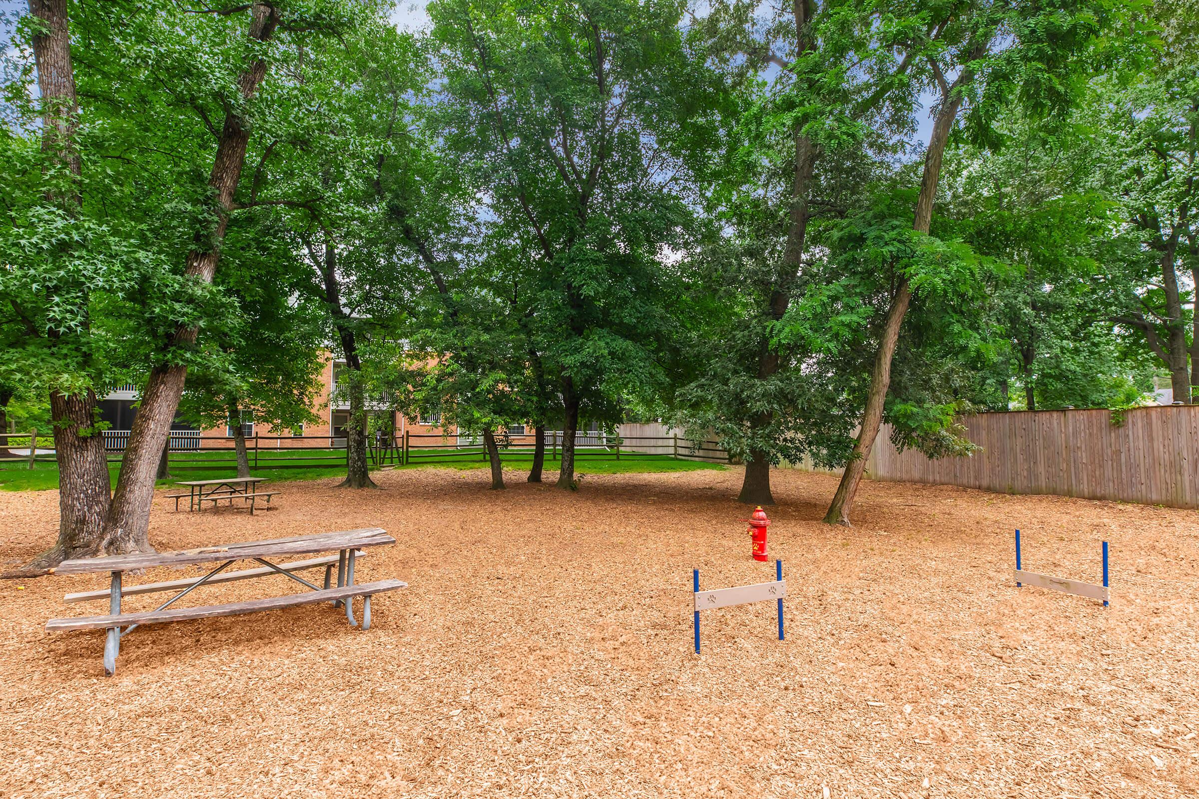 A shaded park area featuring a wooden picnic table surrounded by trees. The ground is covered with wood chips, and there is a red fire hydrant nearby. A wooden fence borders the space, providing a sense of enclosure. Ideal for outdoor relaxation or play.