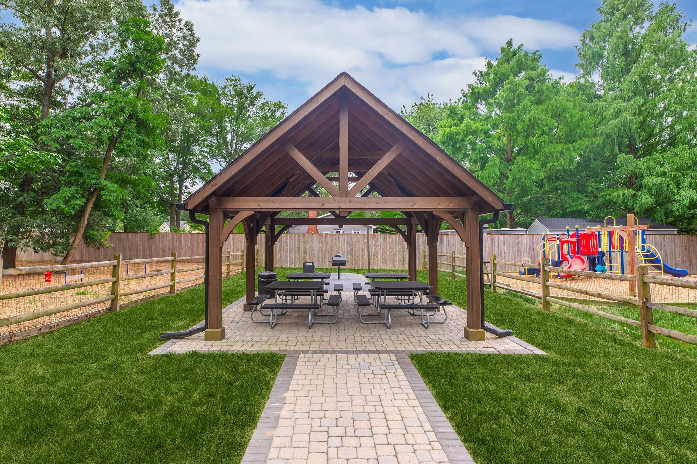 A wooden gazebo with a gabled roof surrounded by green grass and trees. Underneath the gazebo, there are several picnic tables. In the background, there is a playground set and wooden fencing. The sky is partly cloudy, creating a bright outdoor atmosphere.