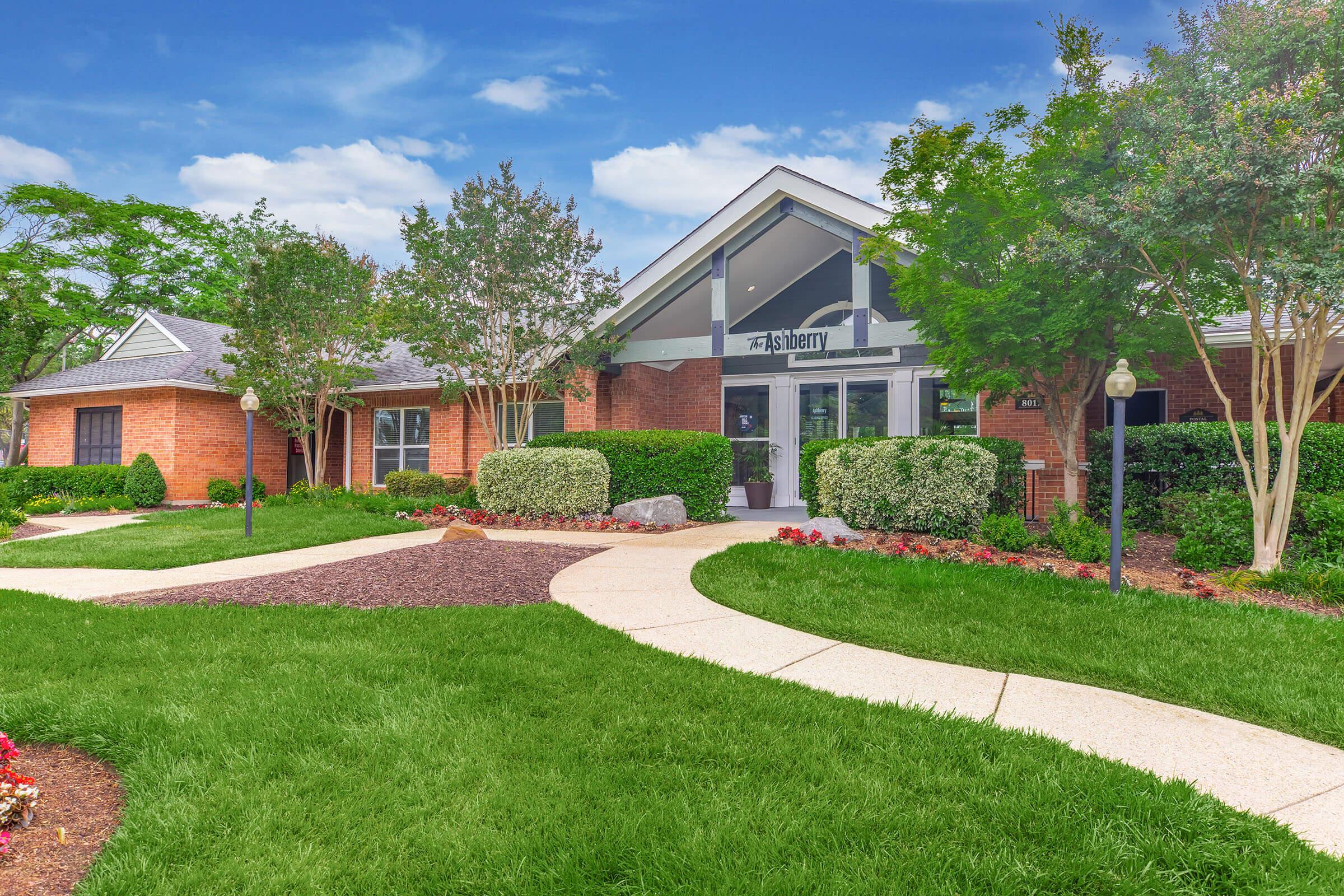 A landscaped exterior view of a brick building with large windows, featuring a modern entrance. The surrounding area includes well-maintained grass, bushes, and flowerbeds, with a walkable path leading to the entrance. The sky is blue with some clouds, creating a pleasant atmosphere.